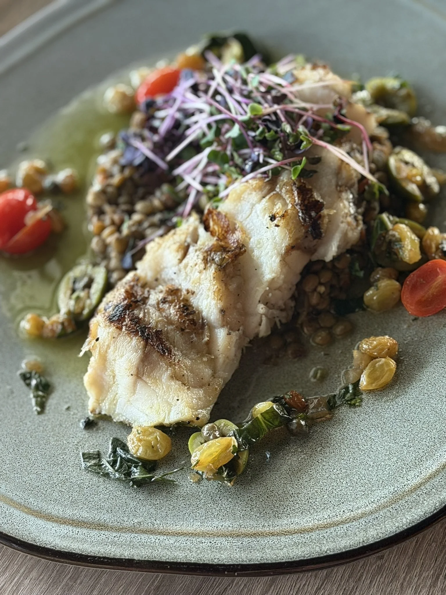 Grilled white fish fillet served on a plate with lentils, cherry tomatoes, sliced jalapeños, and microgreens.