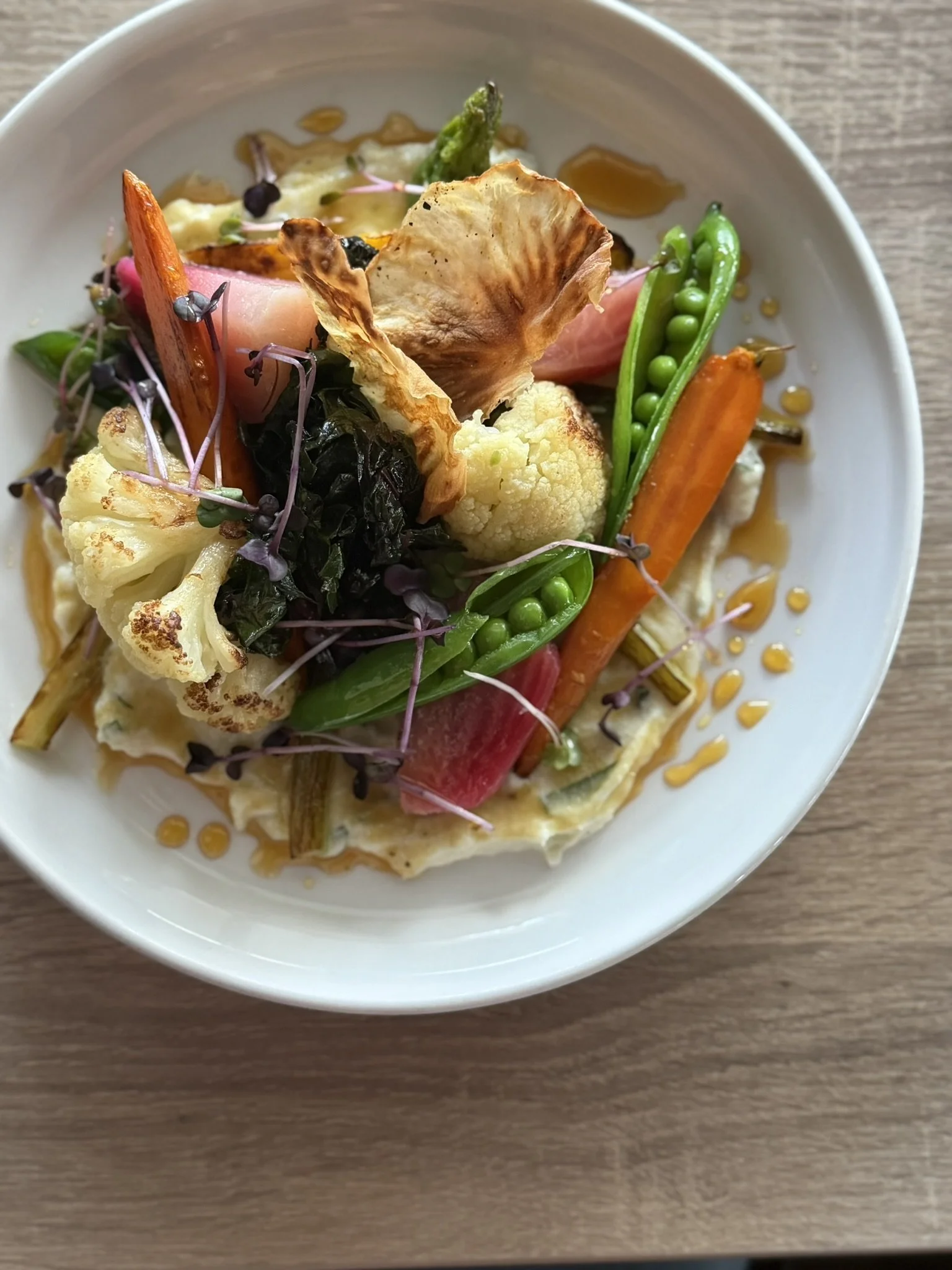 A bowl with assorted roasted vegetables including cauliflower, carrots, snap peas, and purple microgreens, garnished with a sauce or dressing.