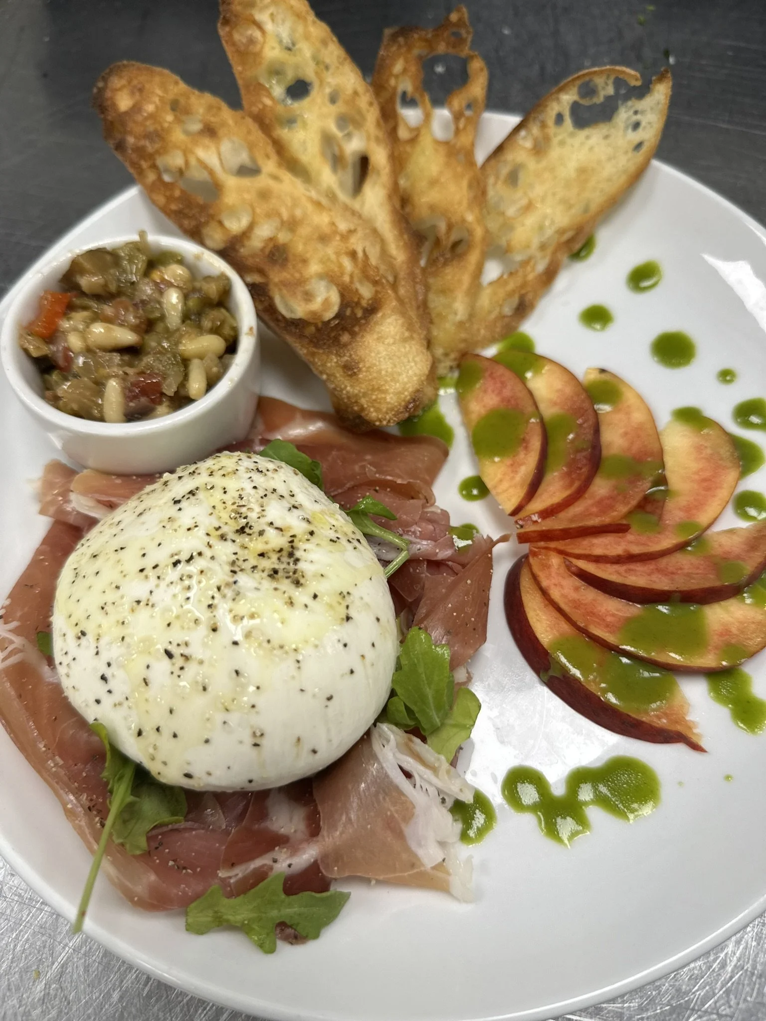 Poached egg with black pepper on top, prosciutto, mixed greens, tomato slices drizzled with green sauce, fried bread chips, and a side of vegetable relish served on a white plate.