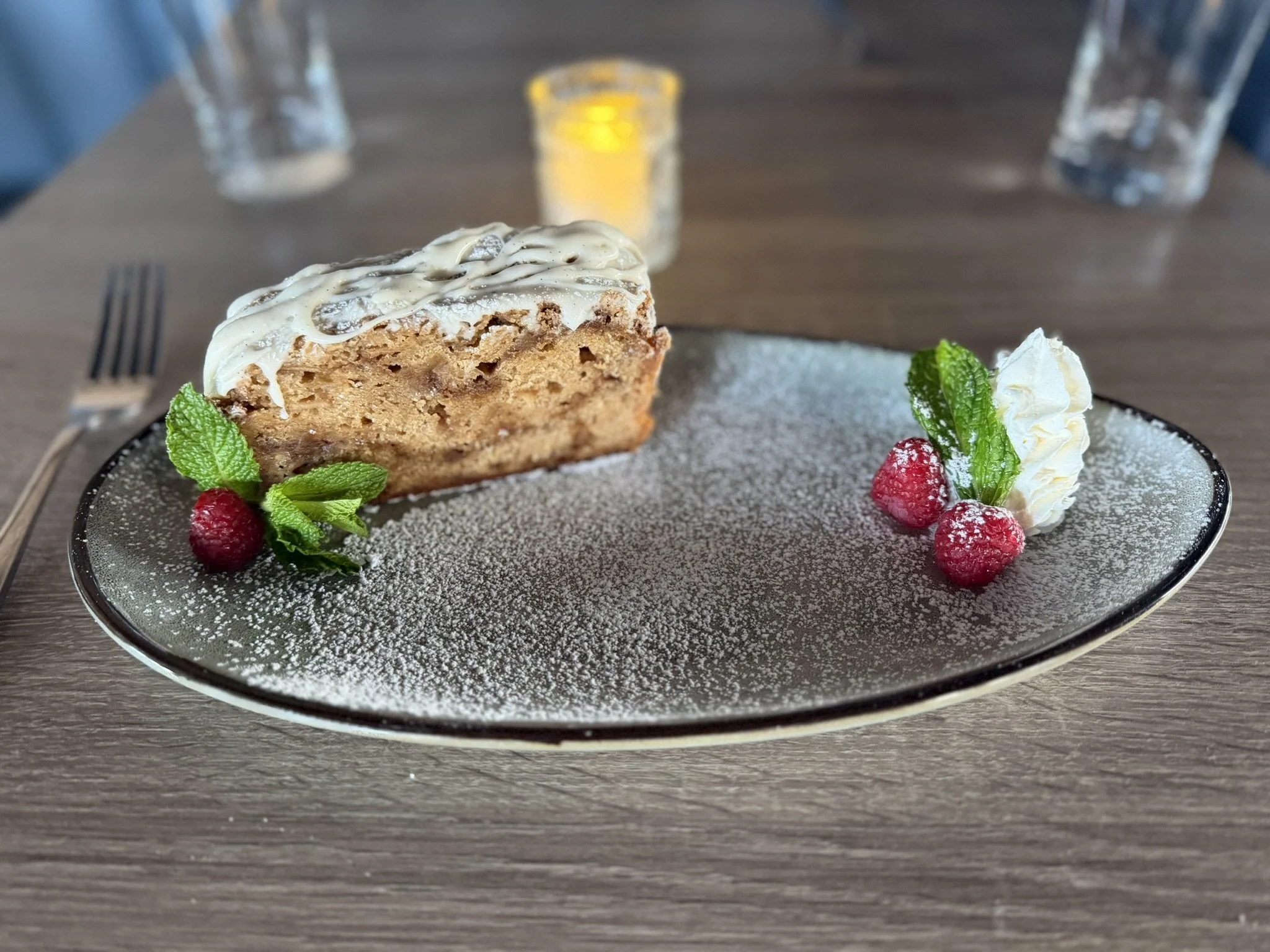 A slice of dessert cake with white icing, garnished with raspberries, mint leaves, and whipped cream on a black-rimmed plate dusted with powdered sugar, set on a wooden table.