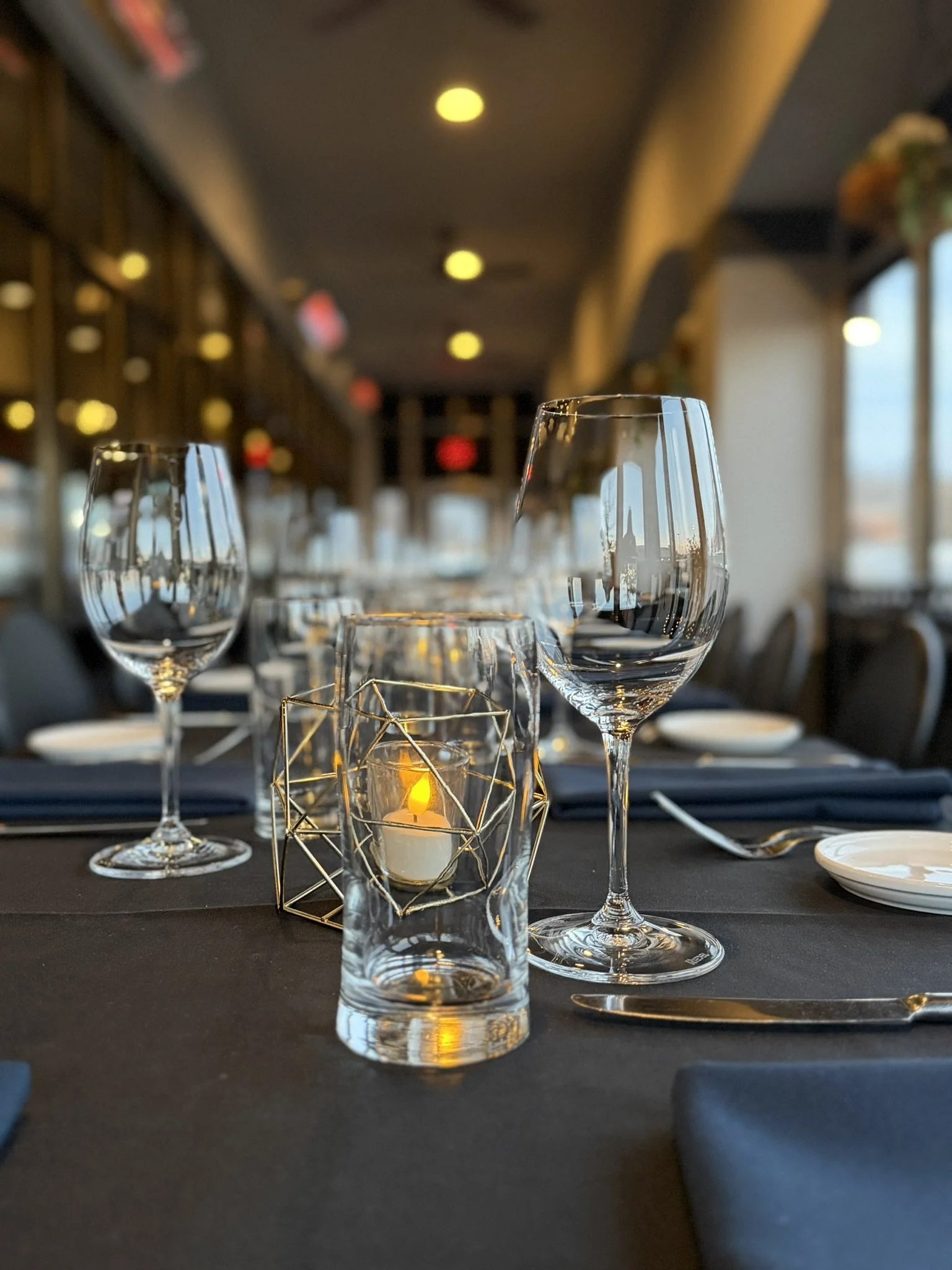 Elegant restaurant table setting with wine glasses, a lit candle in a geometric holder, and a black tablecloth, with a blurred background of a dining area and large windows.