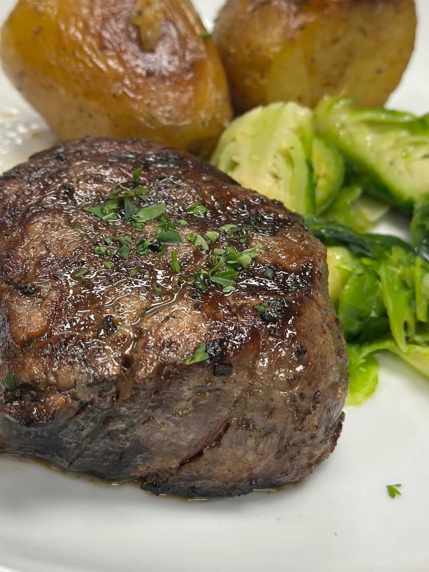 A cooked steak topped with chopped herbs, served with boiled potatoes and Brussels sprouts.
