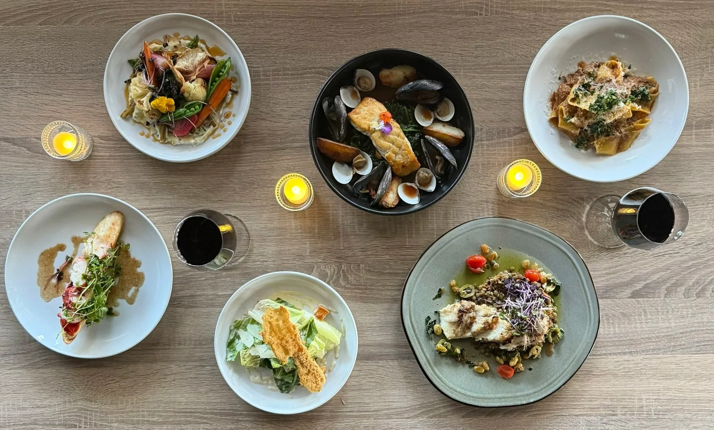 A top-down view of a table with various plates of gourmet food, glasses of red wine, and three lit candles.