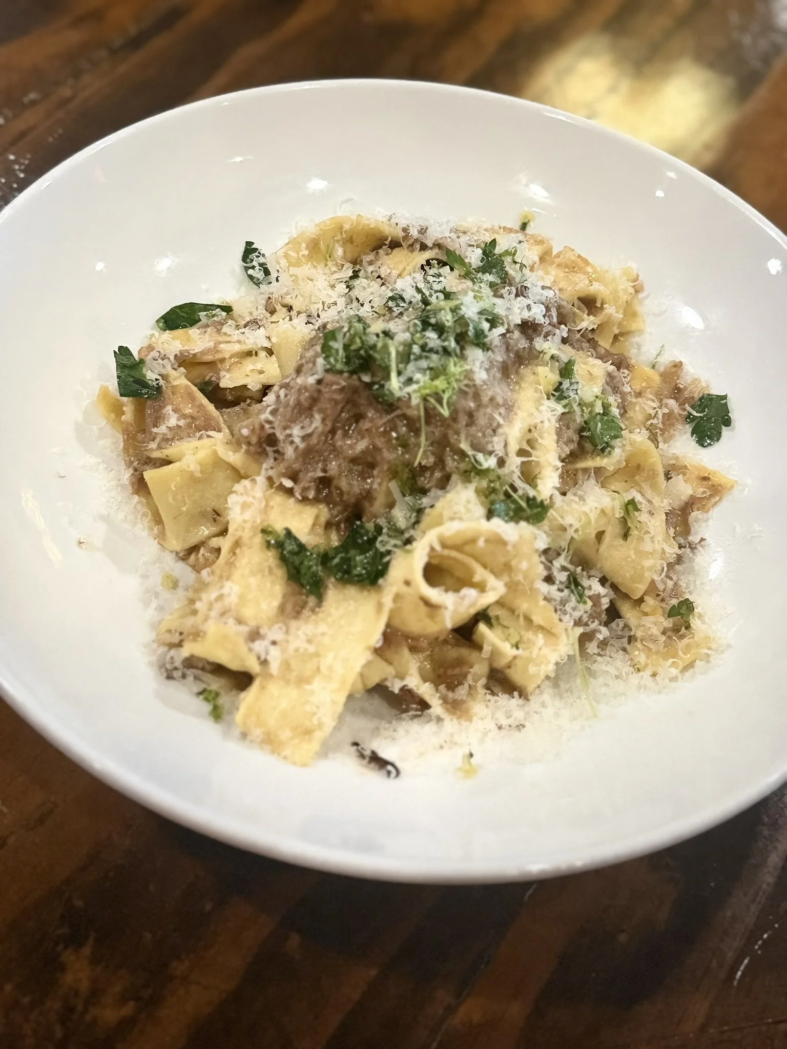 A bowl of pasta with grated cheese, green herbs, and brown sauce on top, on a wooden table.