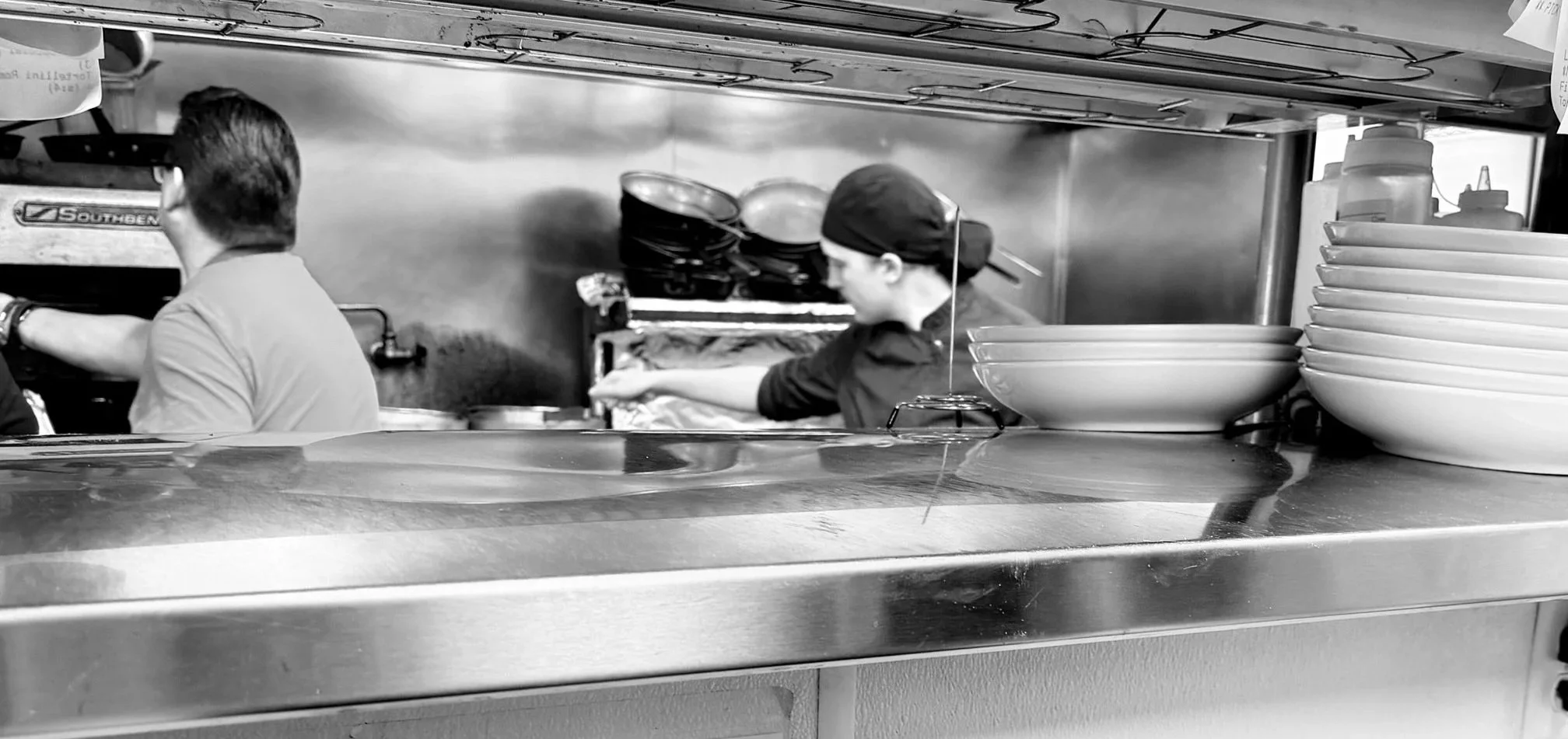 Black and white photo of two people working in a restaurant kitchen, one male and one female, near stacks of bowls and kitchen supplies.