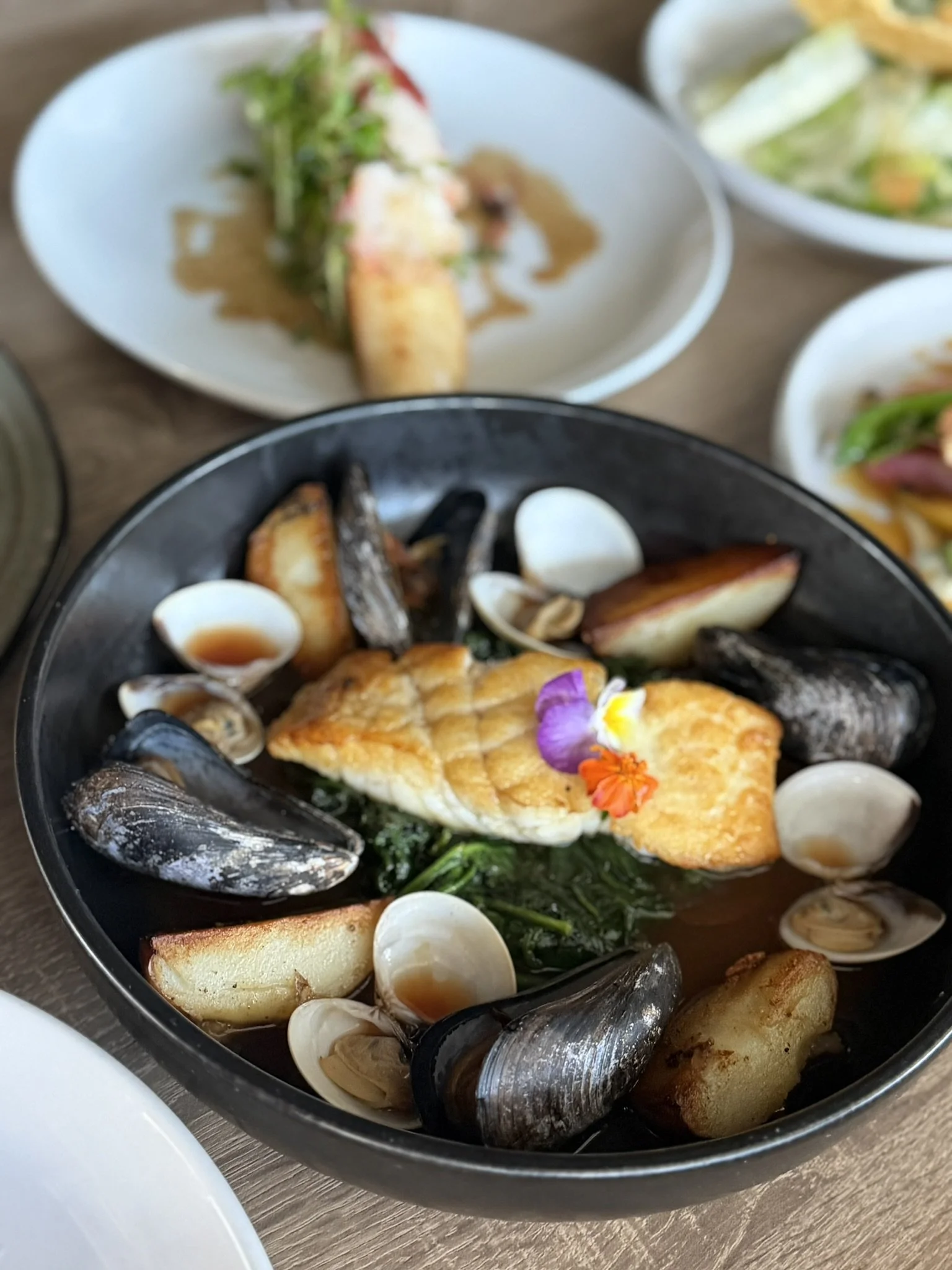 Seafood dish featuring grilled fish, mussels, clams, potatoes, and greens, garnished with edible flowers.
