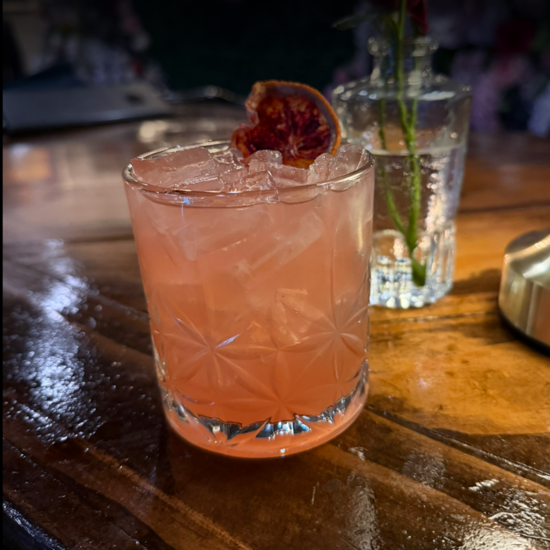 A pink cocktail in a patterned glass garnished with a dried citrus slice, placed on a wooden table with a glass vase of flowers in the background.