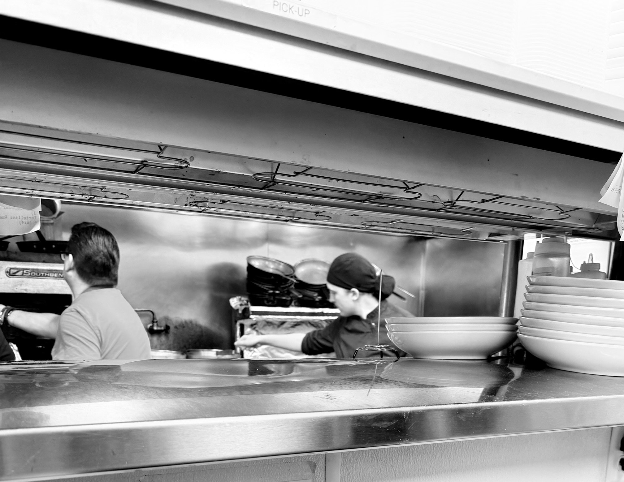 Kitchen scene viewed from behind a counter, showing two employees working in a commercial kitchen with stacked bowls and kitchen equipment.