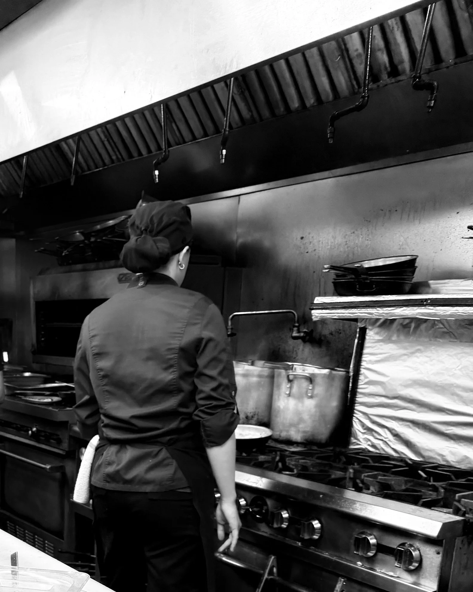 A female chef wearing a chef's hat and uniform, standing in a commercial kitchen, cooking on a stove with large pots and pans.