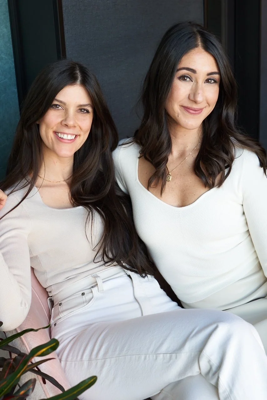 Co-Founders, Elizabeth Wellington and Sarah Battani Sams, sitting close together, wearing white tops, against a dark background.