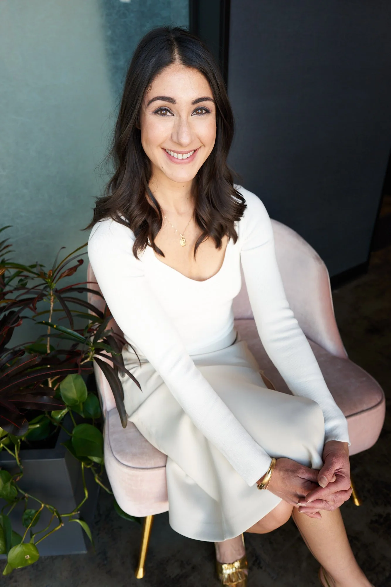 Co-Founder, Elizabeth Wellington, wearing a white top and skirt, sitting on a pink chair next to a green plant. She has shoulder-length dark hair and is wearing jewelry, including a necklace and bracelet.