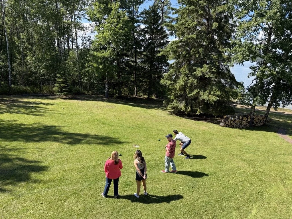 Four people playing lawn games outside on a sunny day with green grass, tall trees, and a forested area in the background.