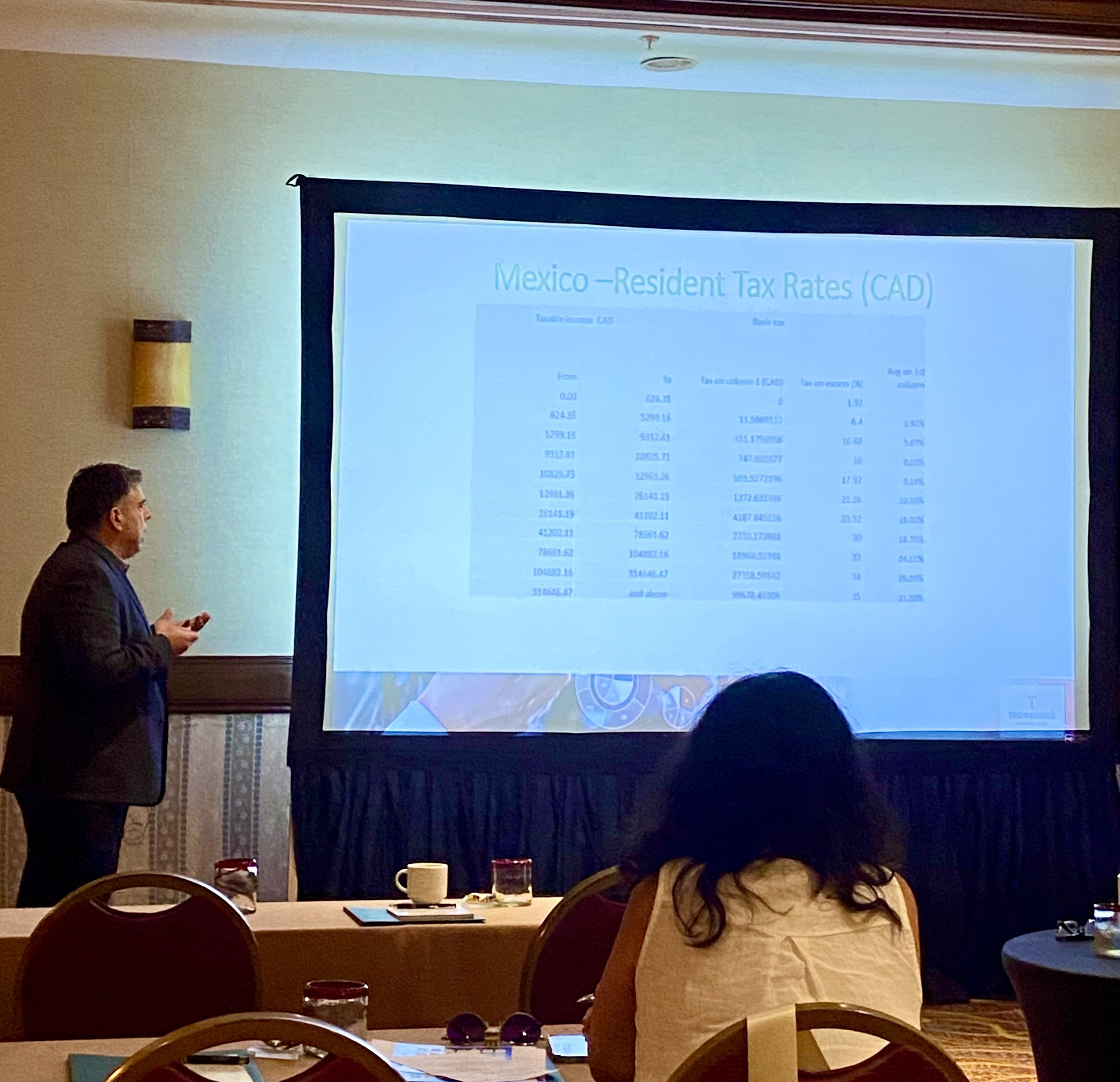 A man in a suit giving a presentation on a large screen with data about Mexico resident tax rates, while a woman with dark hair sits at a table in a conference room.