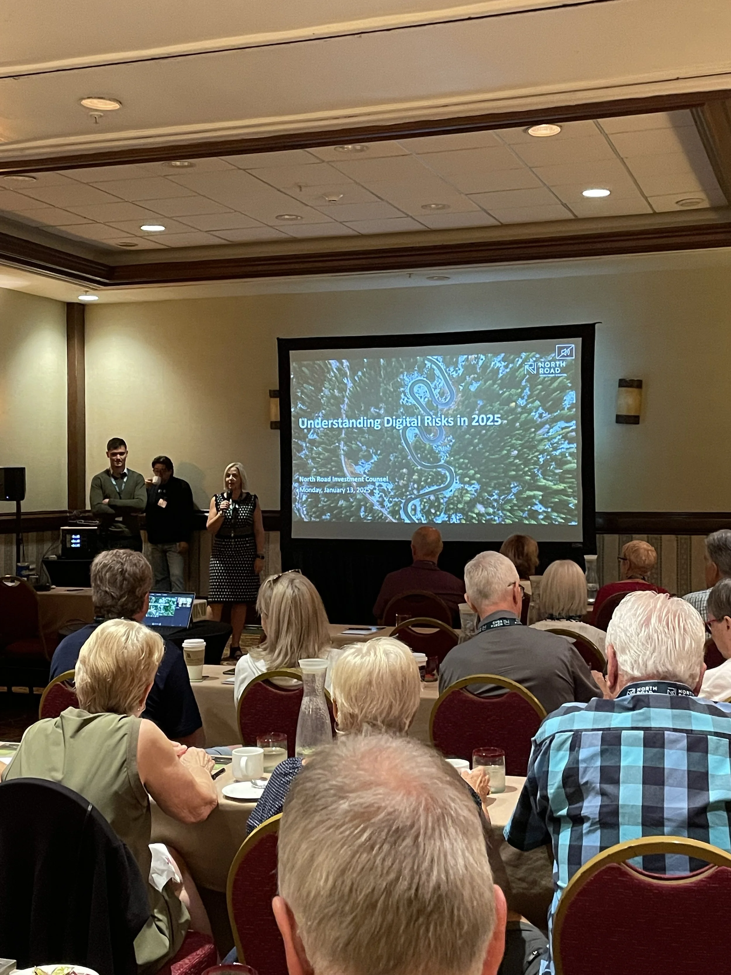 A conference room with attendees listening to a presentation on digital risks in 2025. There are three speakers standing in front of a large screen displaying the presentation title and agenda.