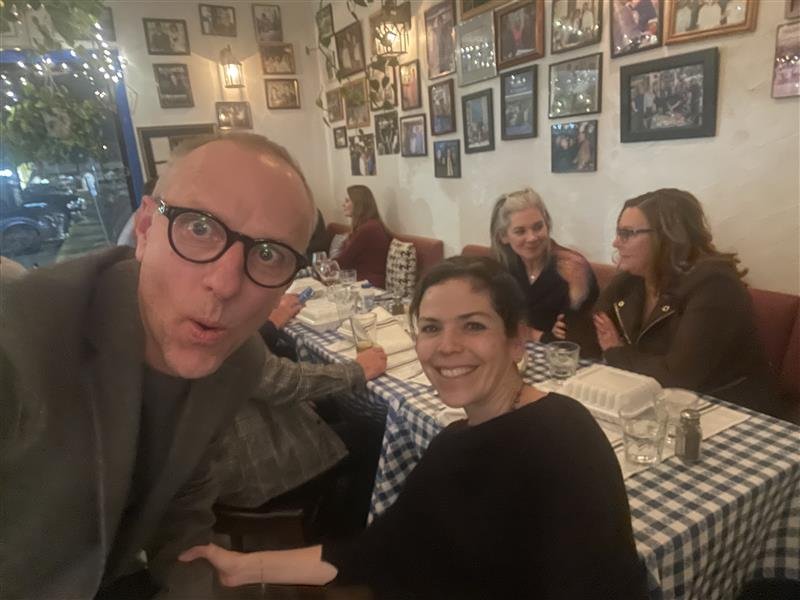 Group of people dining at a restaurant with checkered tablecloths and framed photos on the wall.
