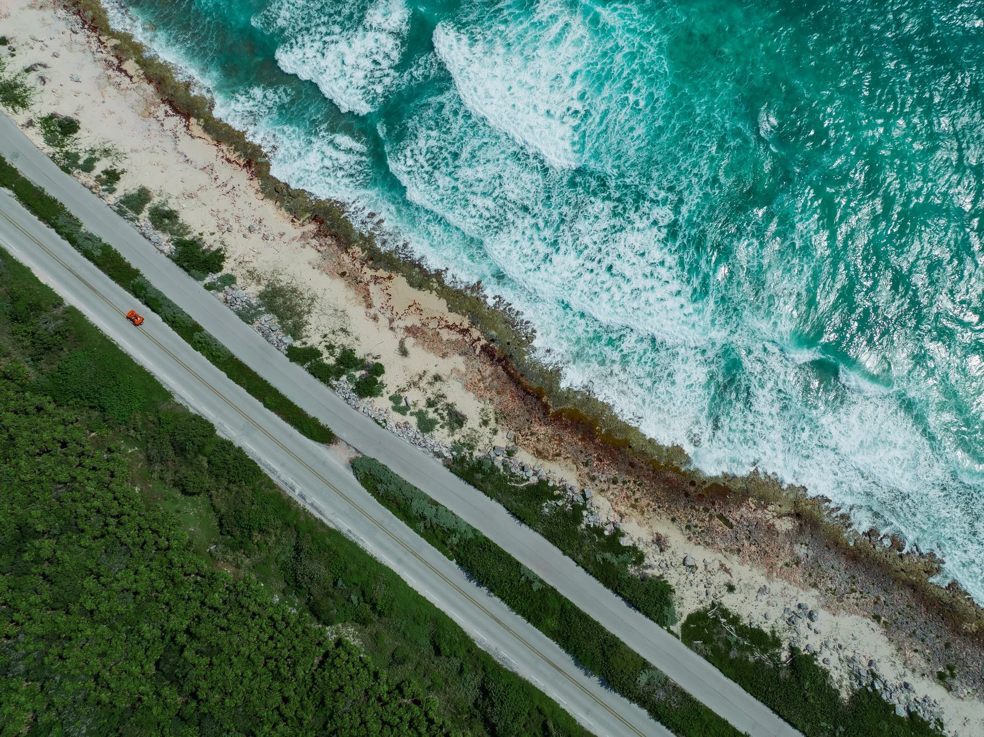 An aerial view of a coastline showing a road running parallel to the ocean, with waves crashing onto the sandy and rocky shore, and lush green trees covering land beside the road.