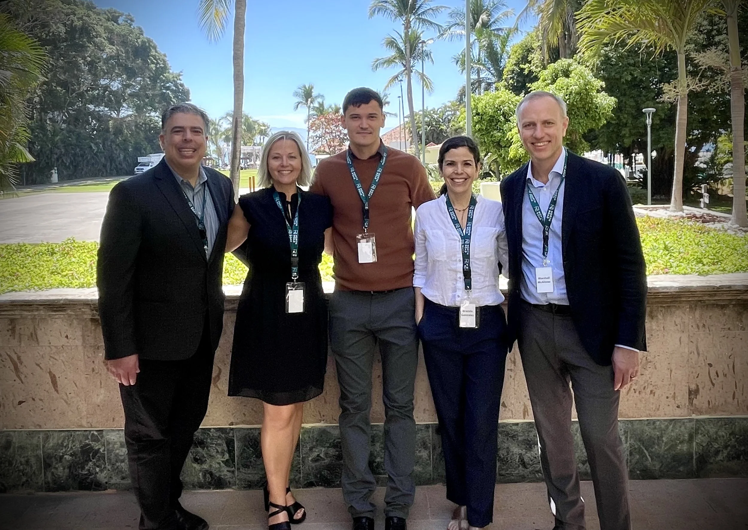 A group of five professionally dressed people standing outside, smiling, with a background of trees, palm trees, and a partly cloudy sky.