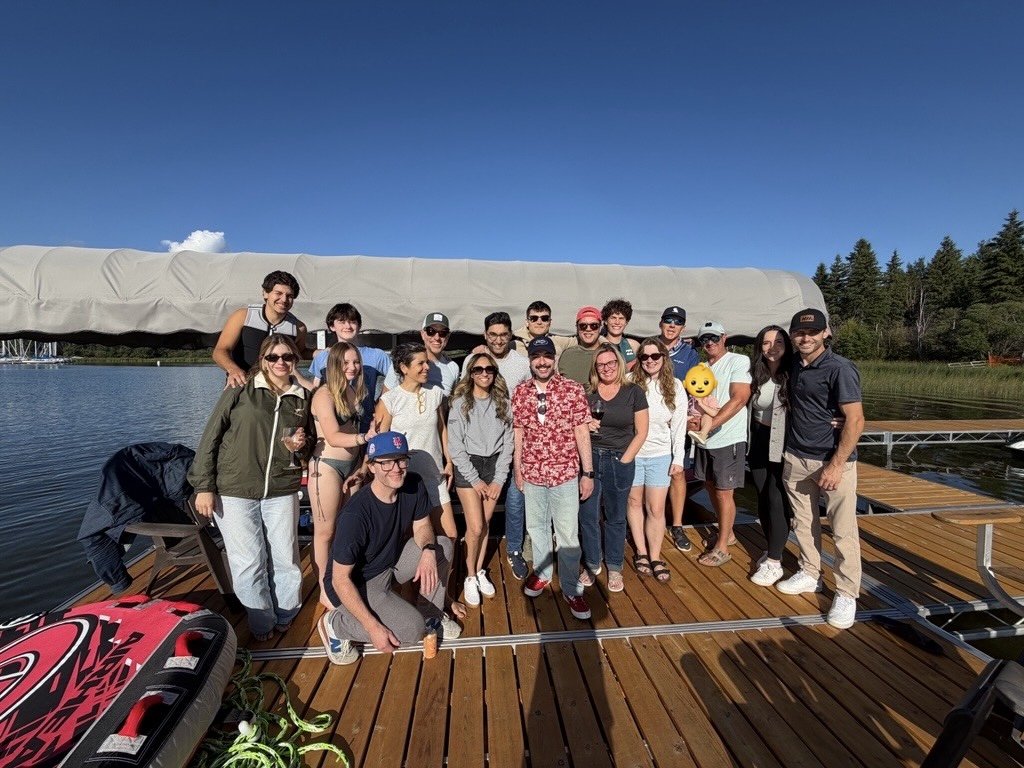 Group of people standing on a wooden dock by a lake with a covered boat, surrounded by trees under a clear blue sky.