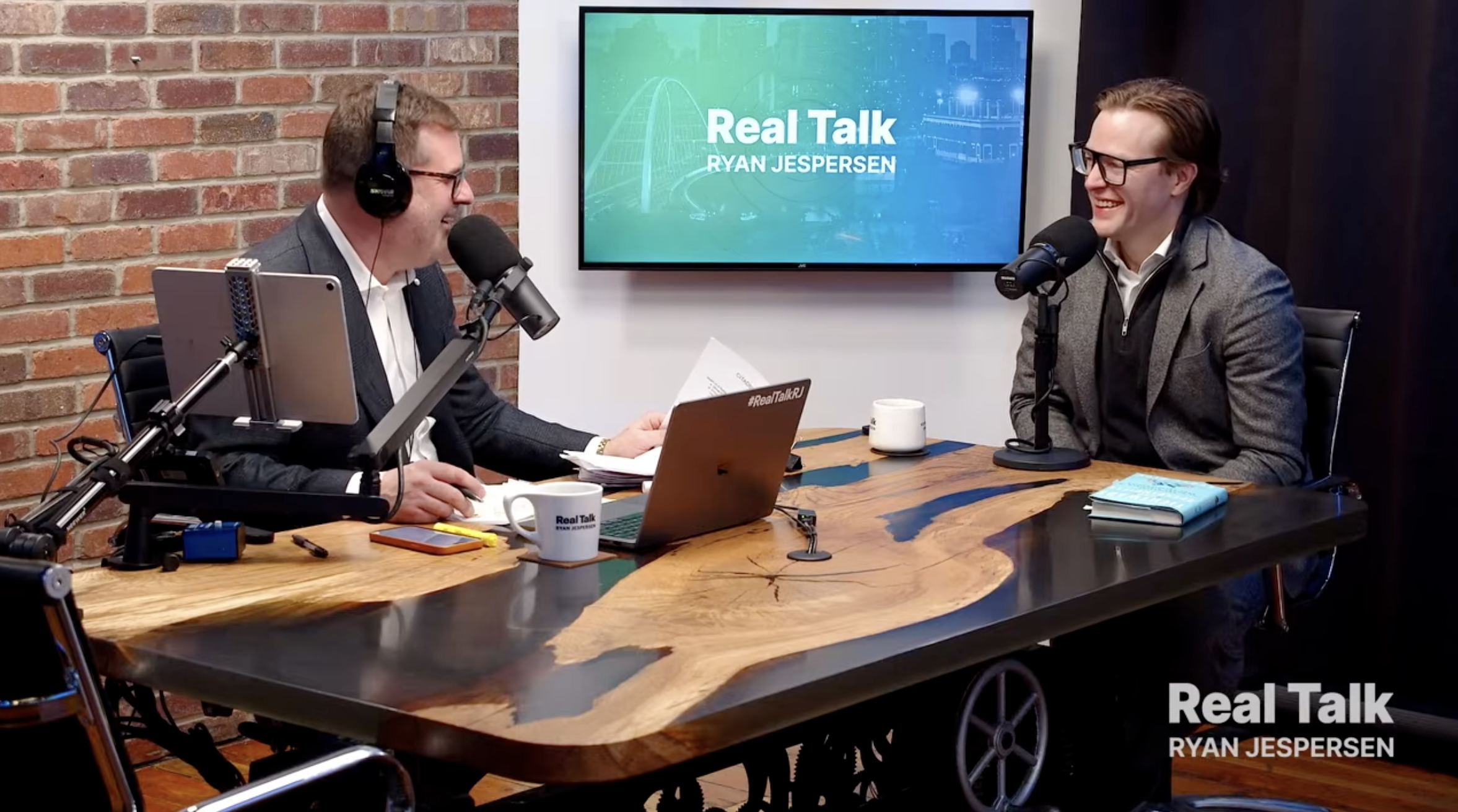 Two men in a podcast studio sitting at a wooden table, wearing headphones and glasses, engaged in conversation. A large screen behind them displays the words 'Real Talk' and 'Ryan Jespersen.' One man has a laptop, a cup, and papers in front of him, while the other has a book and a cup. The studio has a brick wall and dark curtains in the background.