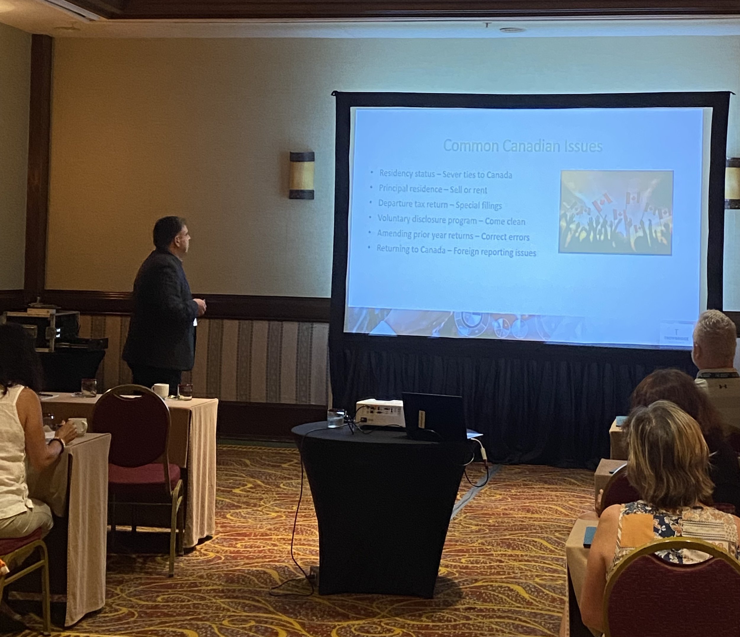 A man in a black suit giving a presentation on common Canadian issues, with an audience seated at round tables in a conference room. The slide on the screen lists issues such as residency status, principal residence, departure tax return, voluntary d