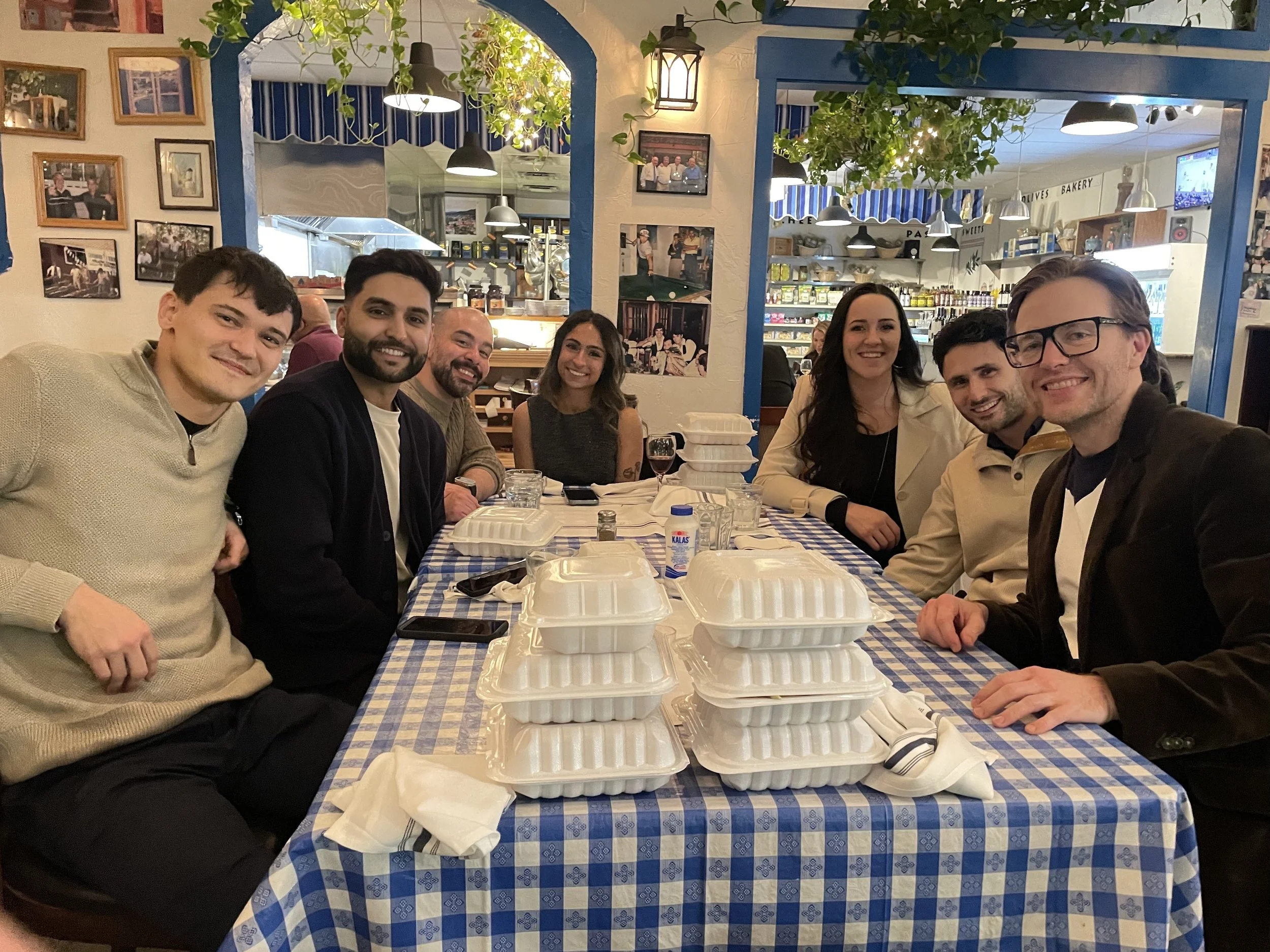 Group of seven friends sitting around a table with stacked takeout containers at a restaurant or cafe, smiling for a photo.