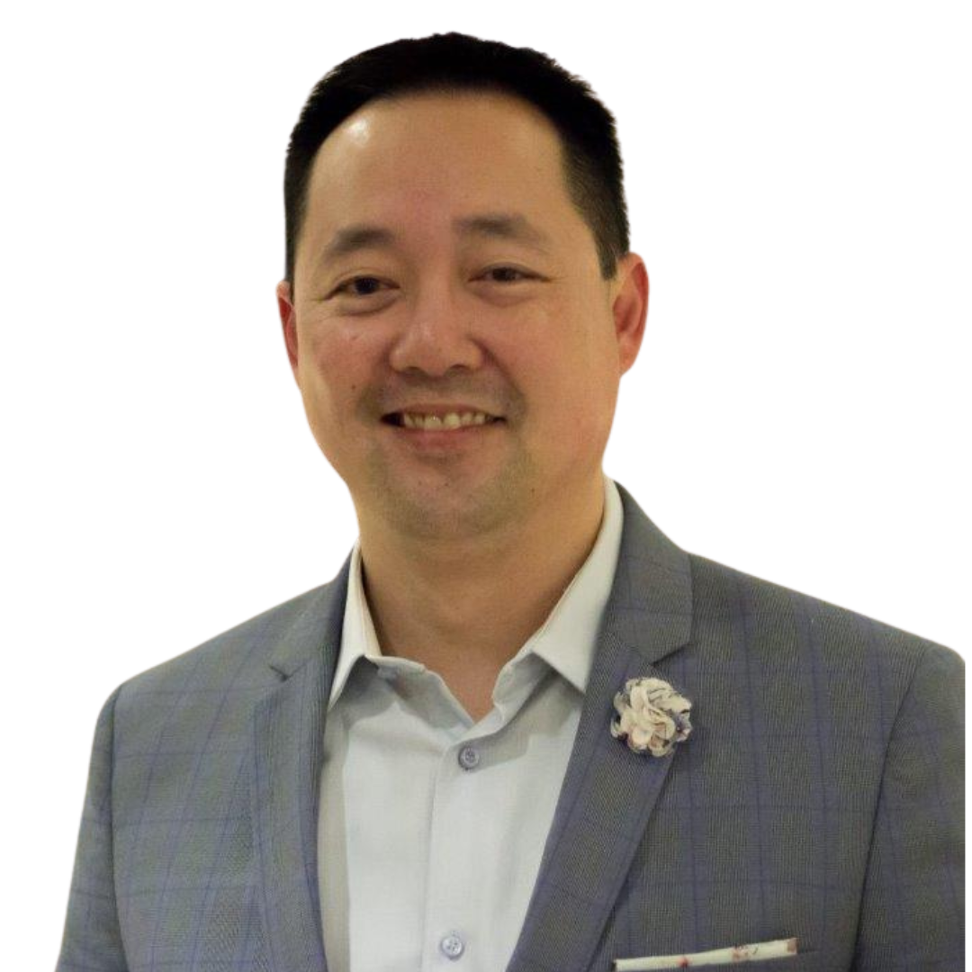 A man in a gray suit and white shirt, smiling, with a flower pin on his lapel, against a black background.
