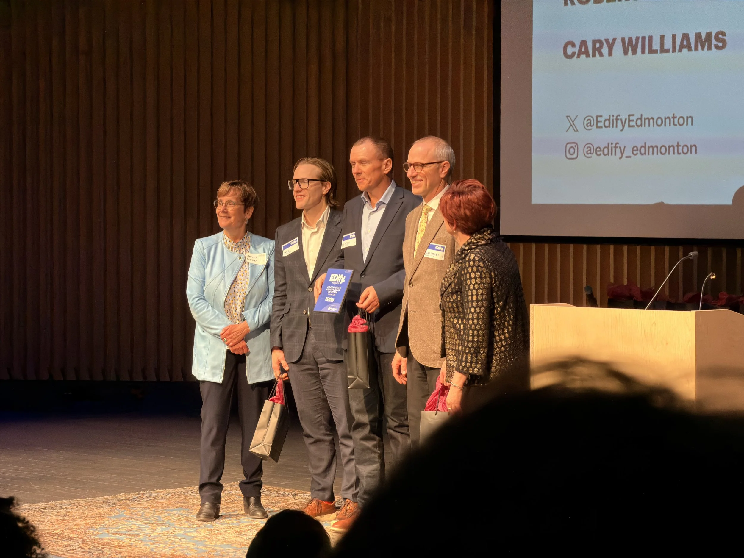 A group of five people standing on a stage in front of a wooden wall, participating in an event. They are all smiling and holding gift bags. The background features a presentation slide with the name 'Cary Williams' and social media handles '@EdifyEd