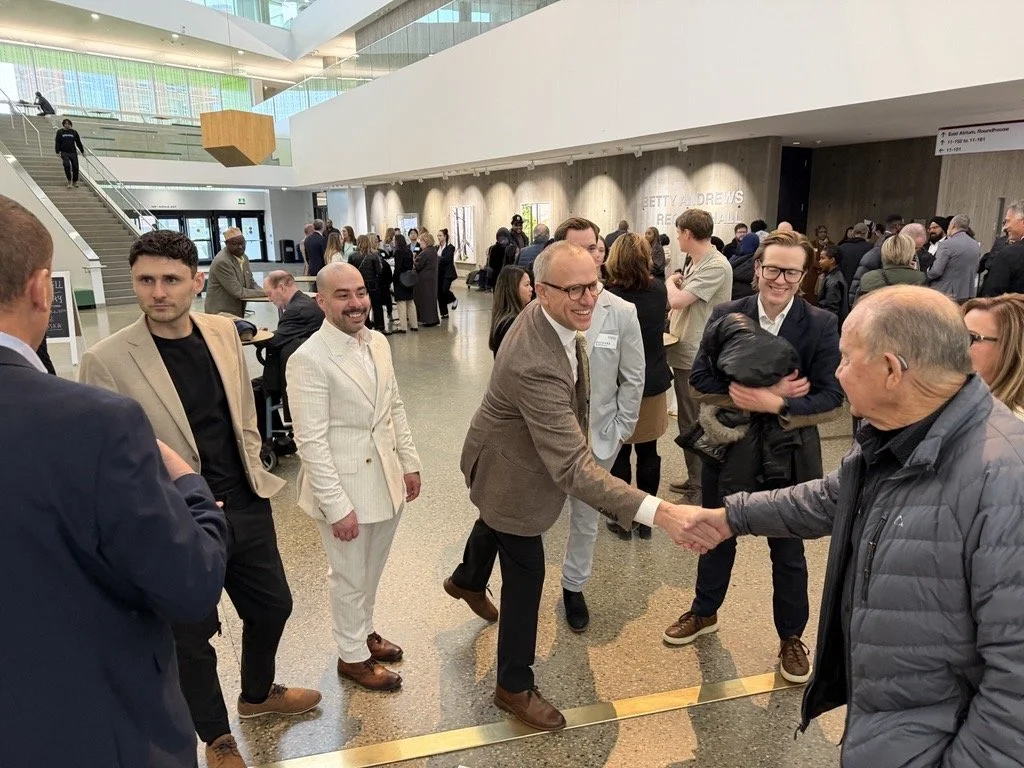 A group of people in a modern lobby engaging in conversation, with some shaking hands, and others standing and talking in the background.