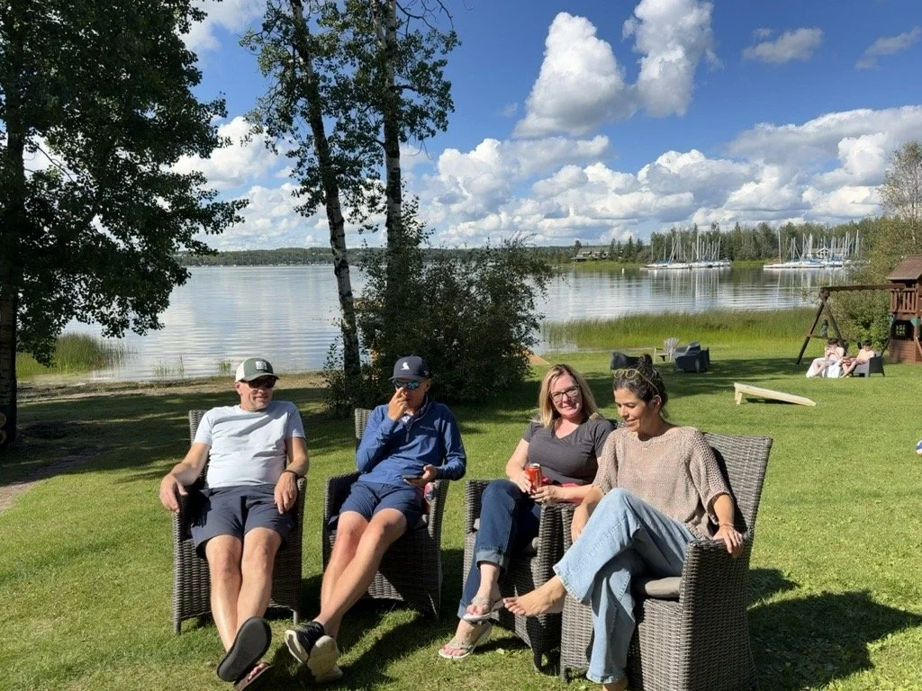 Four friends sitting in outdoor chairs near a lake, with a grassy yard, trees, a small wooden playhouse, and sailboats docked in the water in the background.