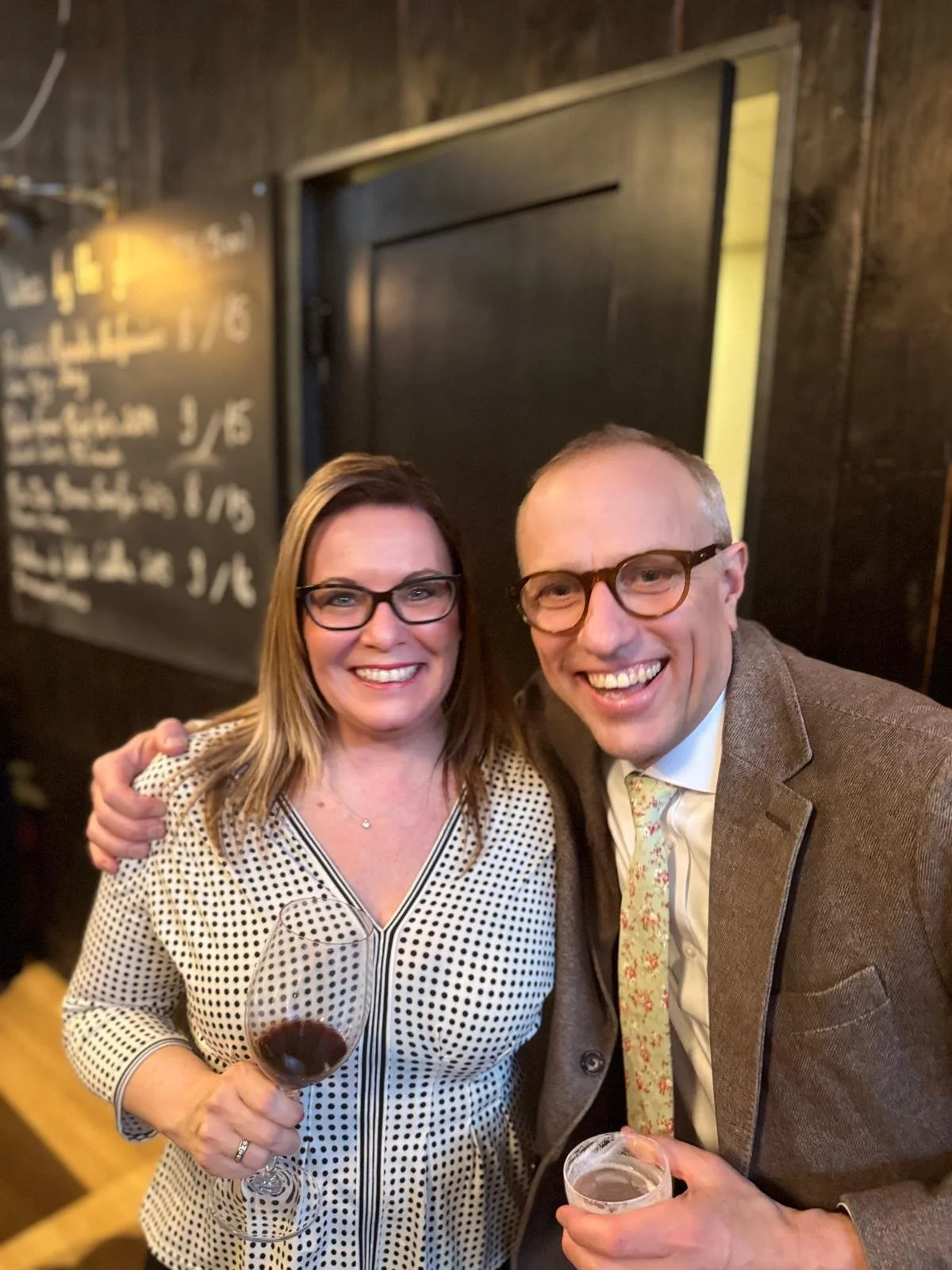 A smiling woman holding a glass of red wine and a smiling man holding a drink in a bar or restaurant setting. They are dressed in semi-formal attire, and there is a blackboard menu in the background.