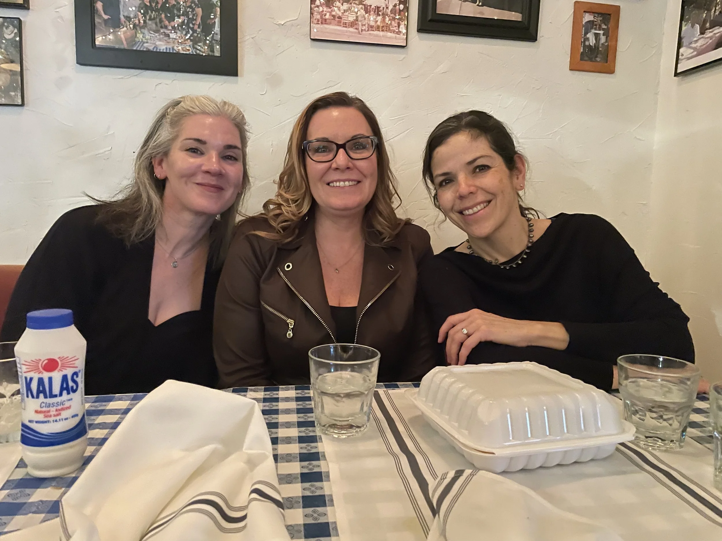 Three women sitting at a restaurant table smiling at the camera, with a takeout container, glasses of water, and a bottle of Kalas yogurt on the table.