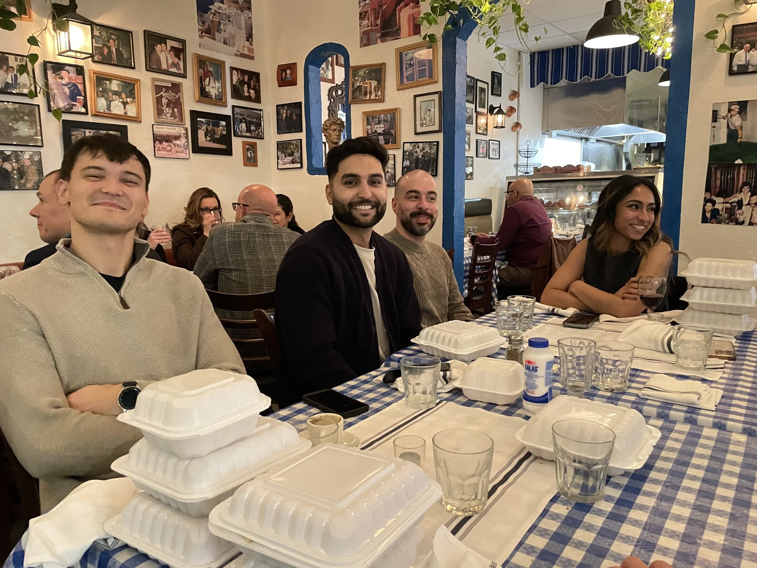A group of four friends sitting at a restaurant table, smiling for the camera. The table has multiple takeout containers, glasses, and utensils. The restaurant interior features framed photos on the wall and a cozy, decorated atmosphere.