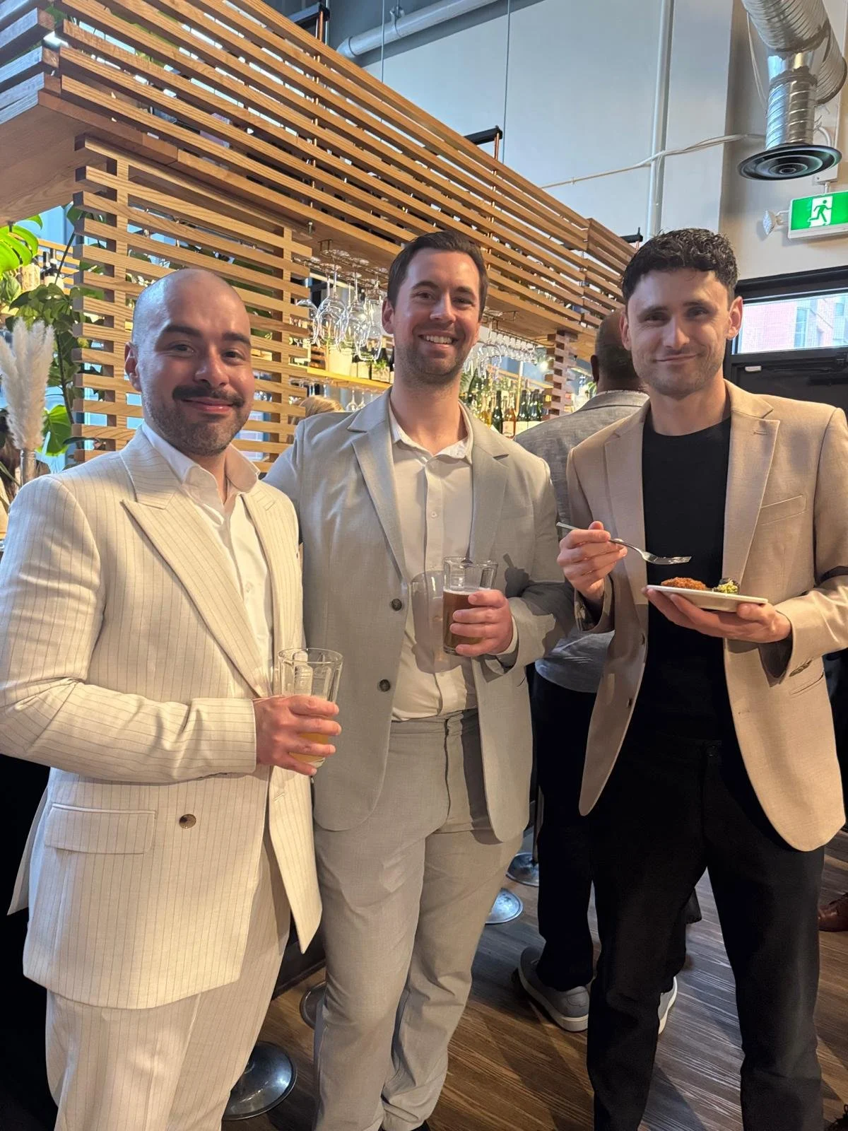 Three men dressed in light-colored suits are standing together at a social event, smiling and holding drinks and a plate of food.