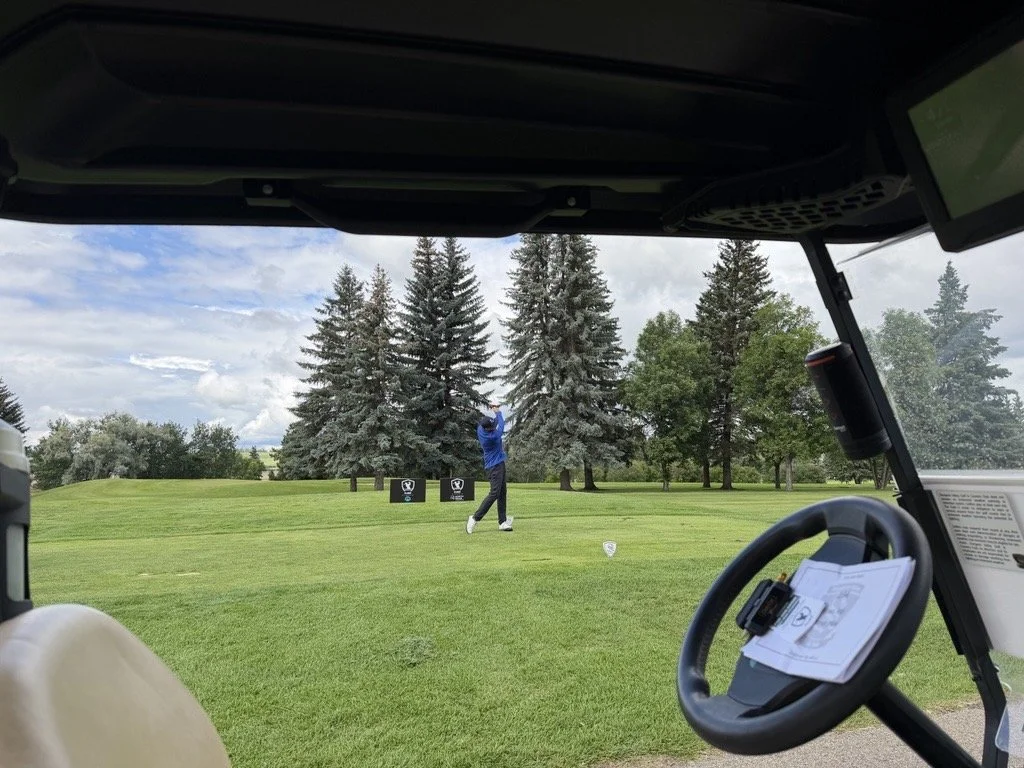 A golfer taking a shot on a golf course, as seen from the perspective inside a golf cart, with trees and a cloudy sky in the background.