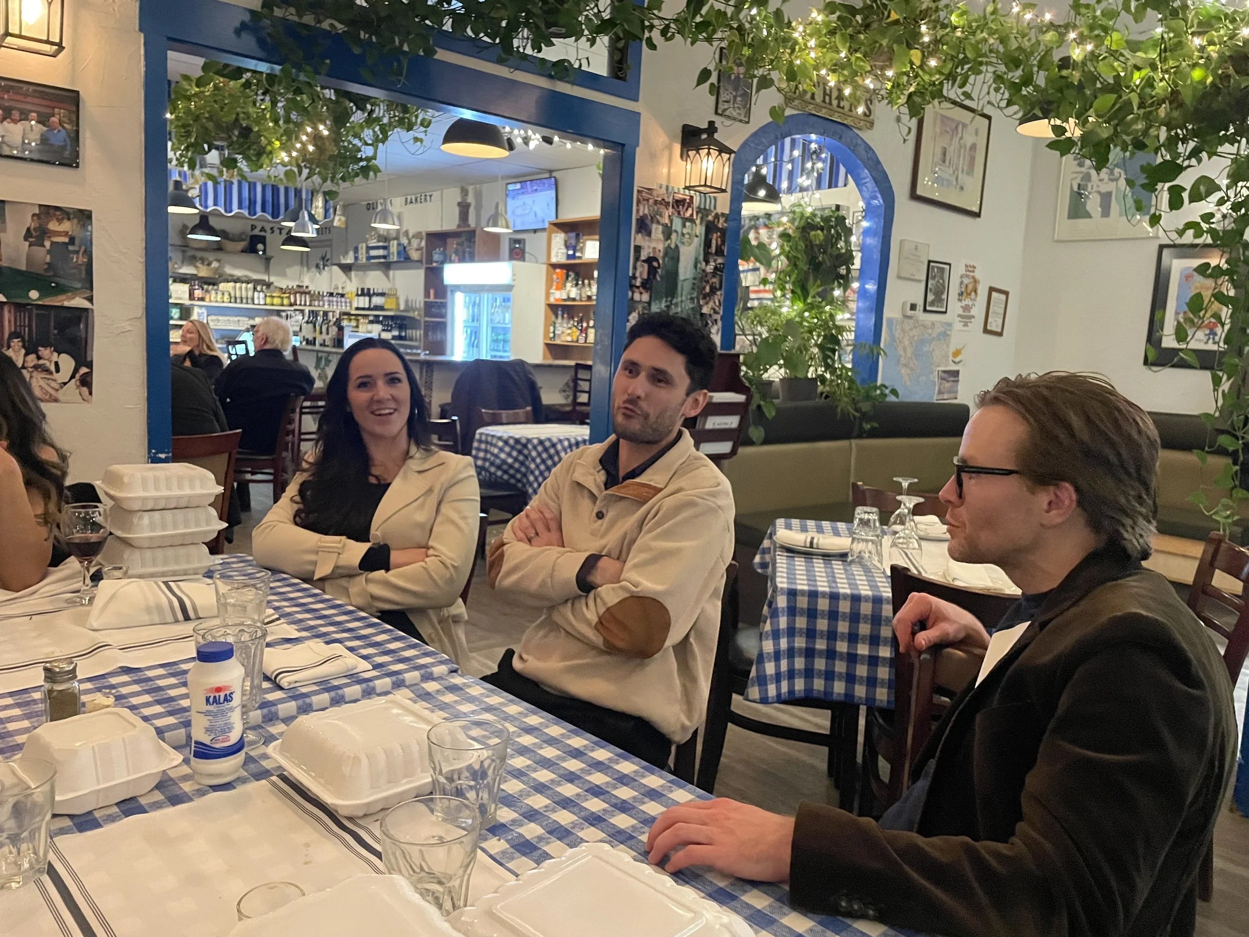 People sitting at a table in a cozy, decorated restaurant with checkered tablecloths, surrounded by plants and framed pictures on the walls, engaging in conversation.
