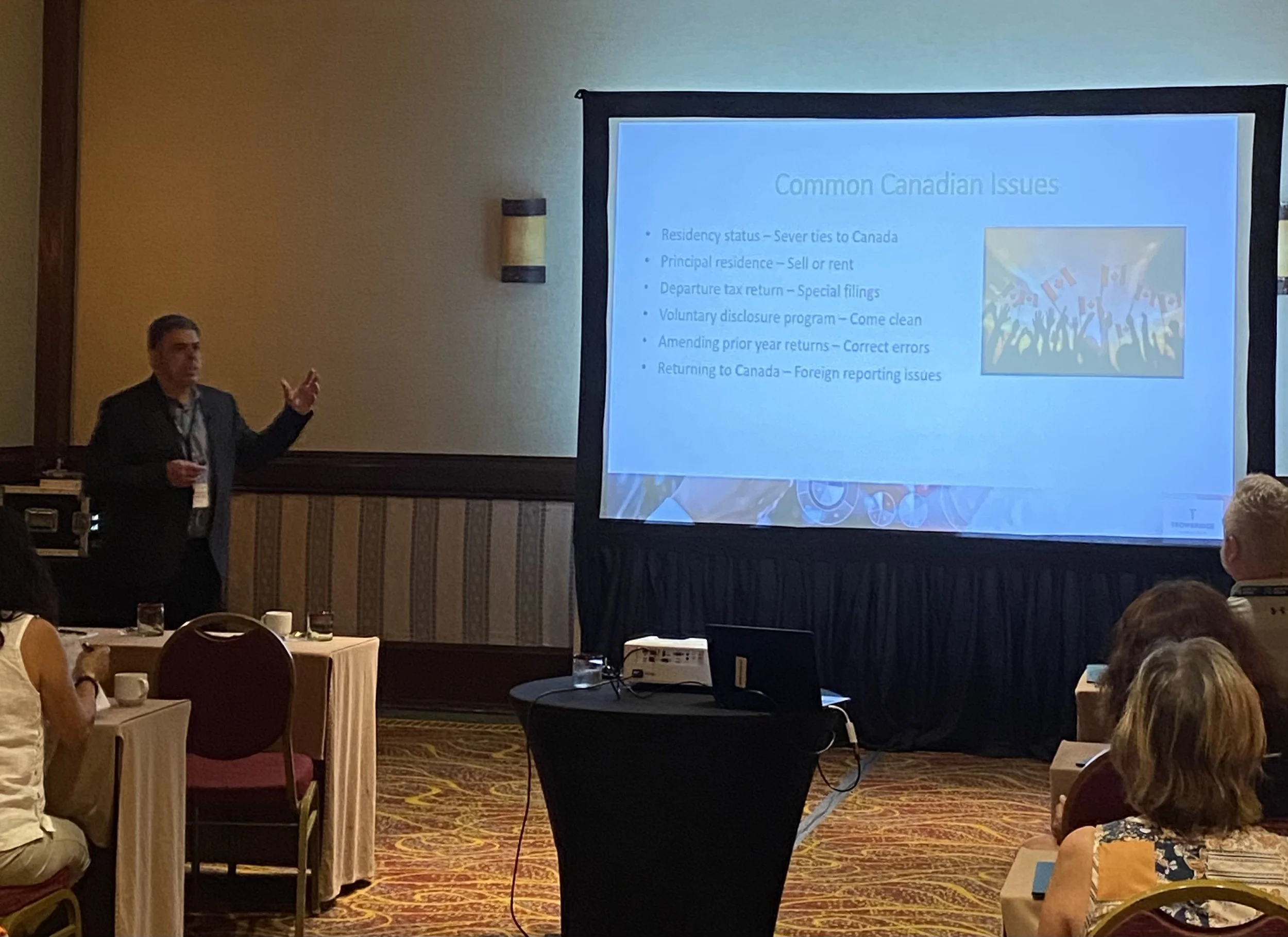A man in a dark suit giving a presentation in a conference room with a slide titled 'Common Canadian Issues' projected on a screen.