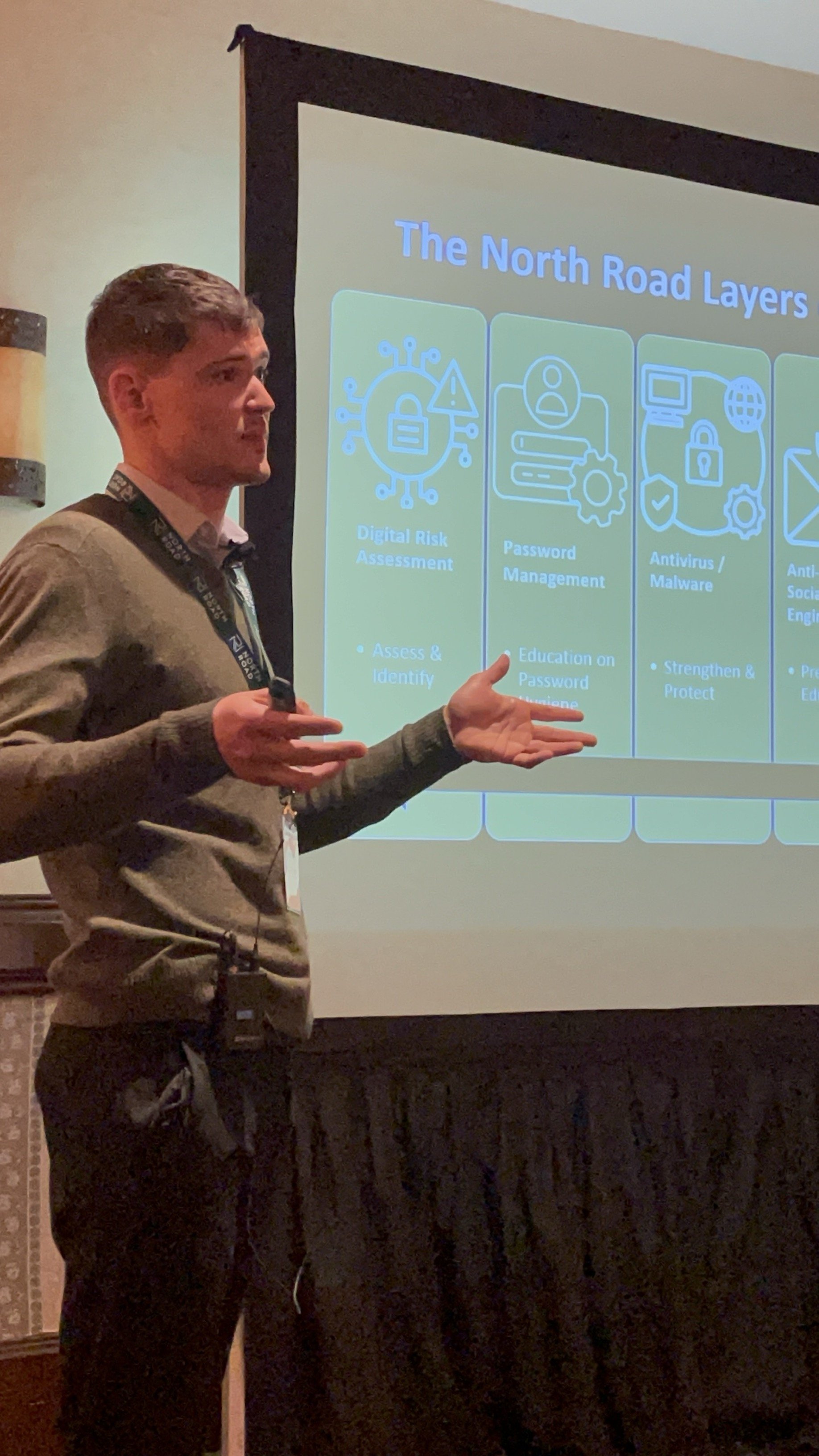 A man giving a presentation on cybersecurity layers, with a slide titled 'The North Road Layers' displayed behind him. The slide includes icons and points about digital risk assessment, password management, antivirus/malware, and social engineering.
