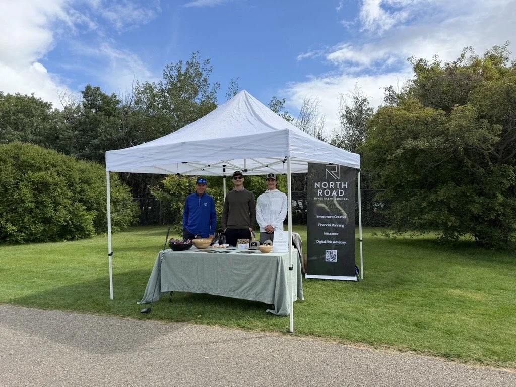 Three men standing behind a table under a white canopy tent outdoors, near a sign for North Road Investment Counsel, on a grassy area with trees and a partly cloudy sky in the background.