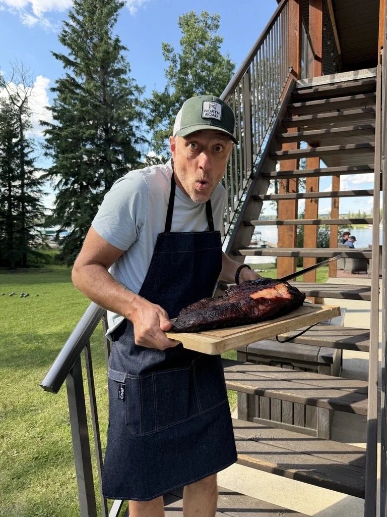 A man standing outdoors on a wooden staircase, holding a large smoked or cooked meat on a cutting board, making a surprised face. He is wearing a gray t-shirt, a cap with a "North Road" logo, and an apron. The background shows green grass, trees, and