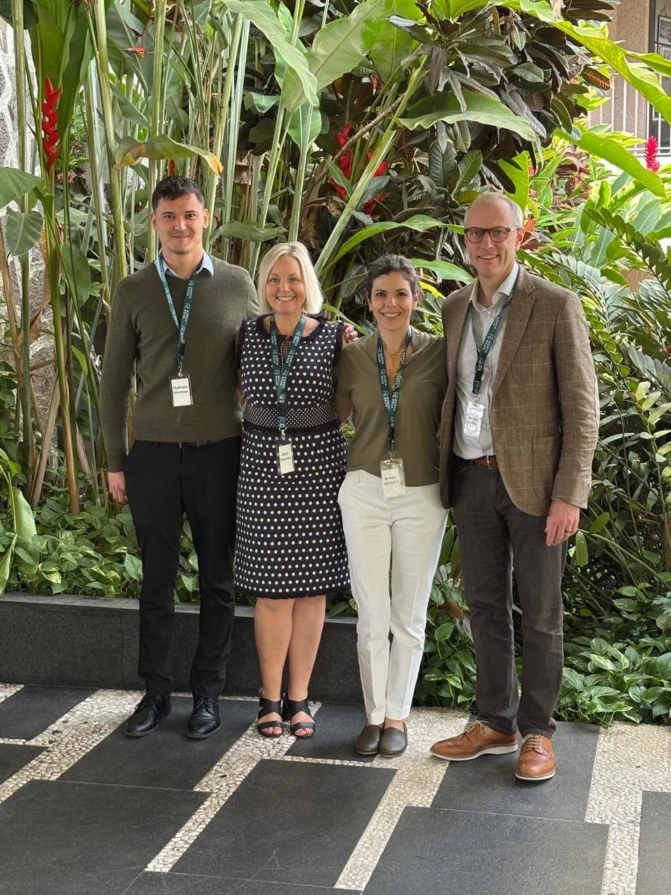 Four people standing side by side in front of lush green plants, all wearing conference badges and lanyards.
