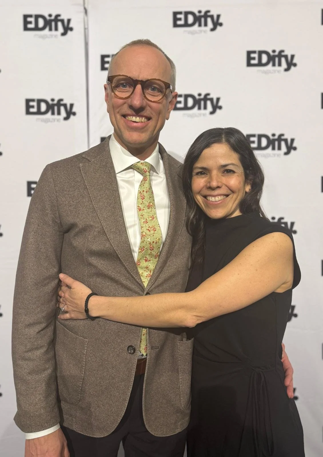 A man and woman smiling and hugging at a professional event with a step and repeat backdrop that says 'EDify magazine'.