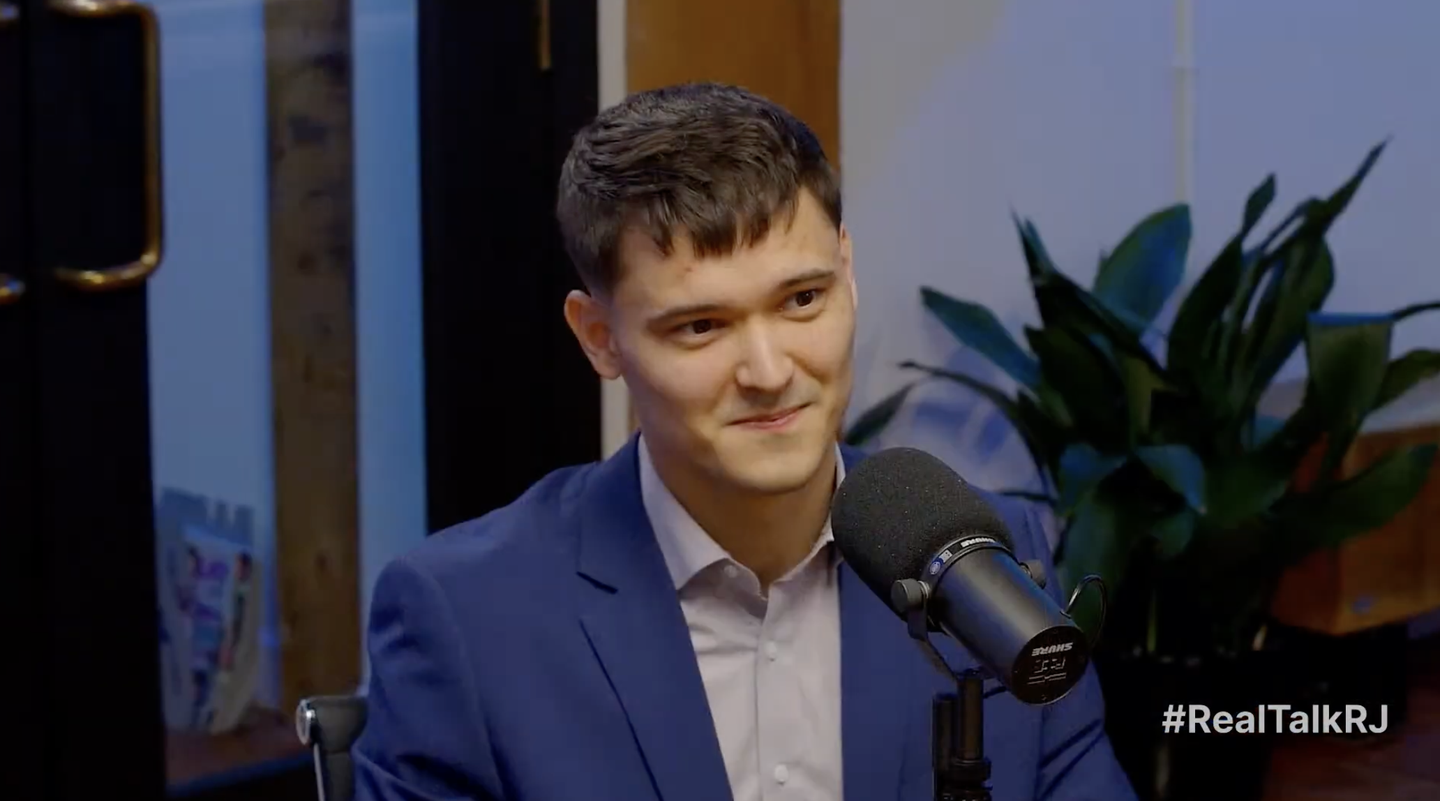 A young man in a blue suit and white shirt is sitting at a desk, speaking into a microphone and smiling. There is a large plant behind him and a bookshelf or cabinet in the background. The hashtag #RealTalkRJ is visible in the lower right corner.