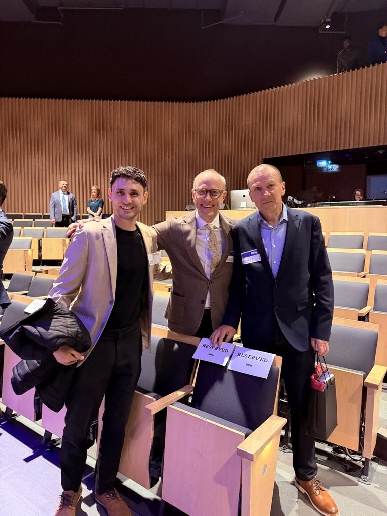 Three men standing in an auditorium, smiling and posing for a photo, with reserved signs on the chairs in front of them.