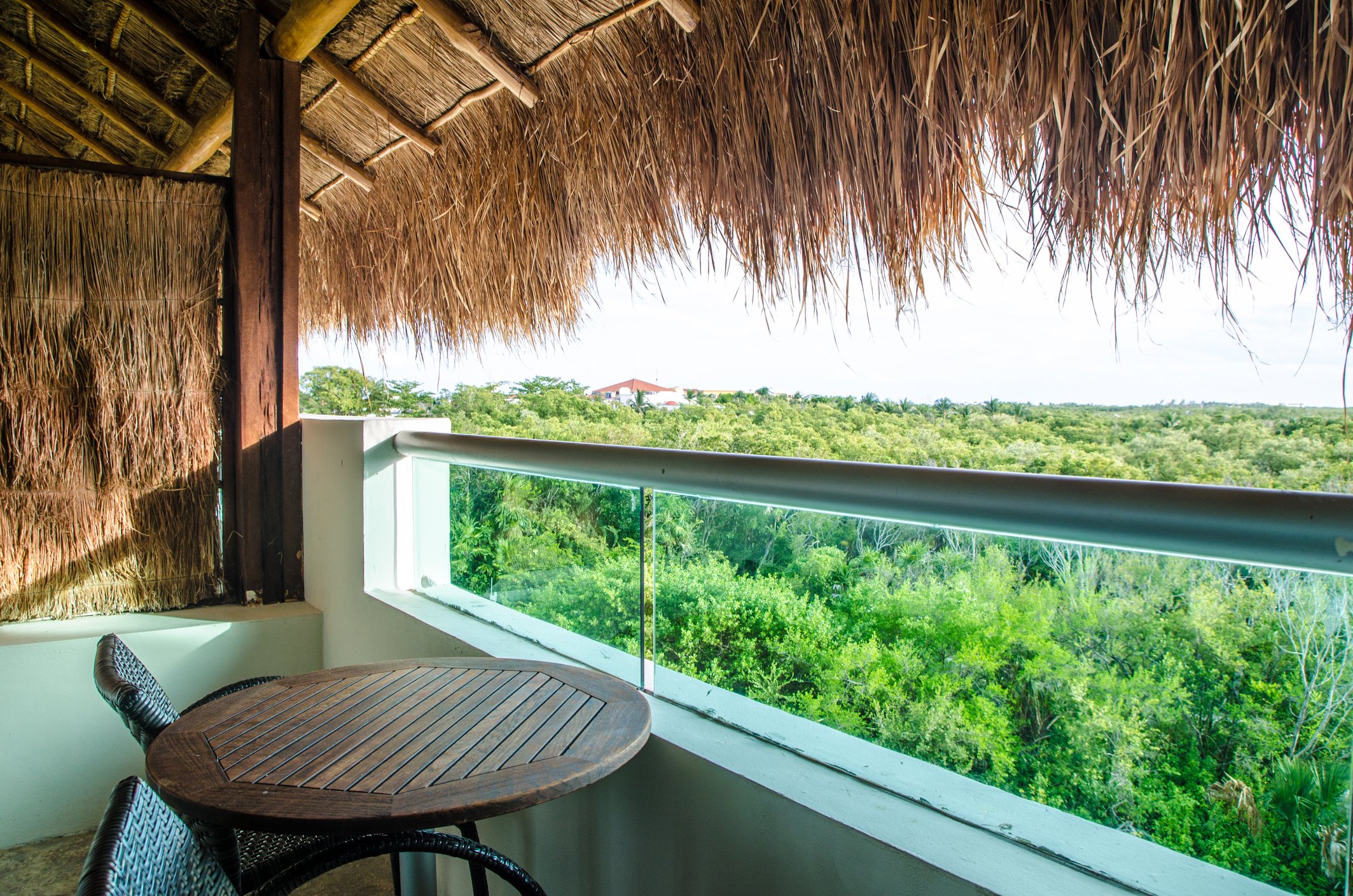 A balcony with a thatched roof overhang, overlooking a lush green forest landscape under a cloudy sky, with a small round wooden table and wicker chairs.