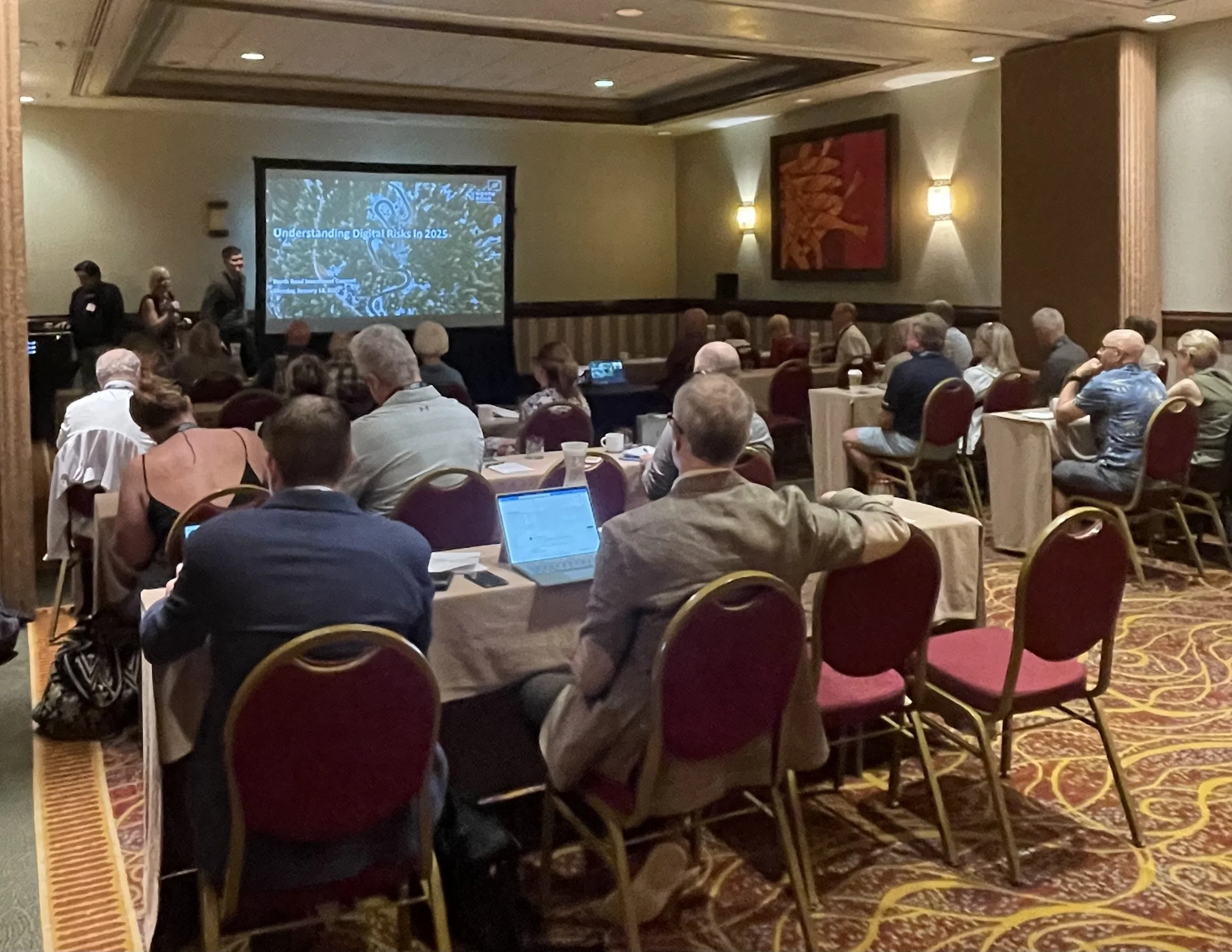 People attending a conference or seminar in a hotel conference room, seated at tables watching a presentation on a large screen.