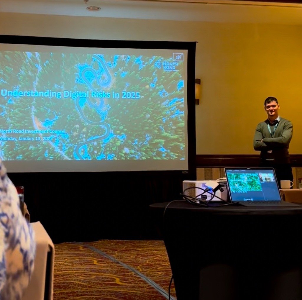 A man standing with arms crossed next to a presentation screen displaying the title 'Understanding Digital Risks in 2025', the date January 13, 2023, and logos for North Road Investment Council and North Road. The presentation appears to be taking pl