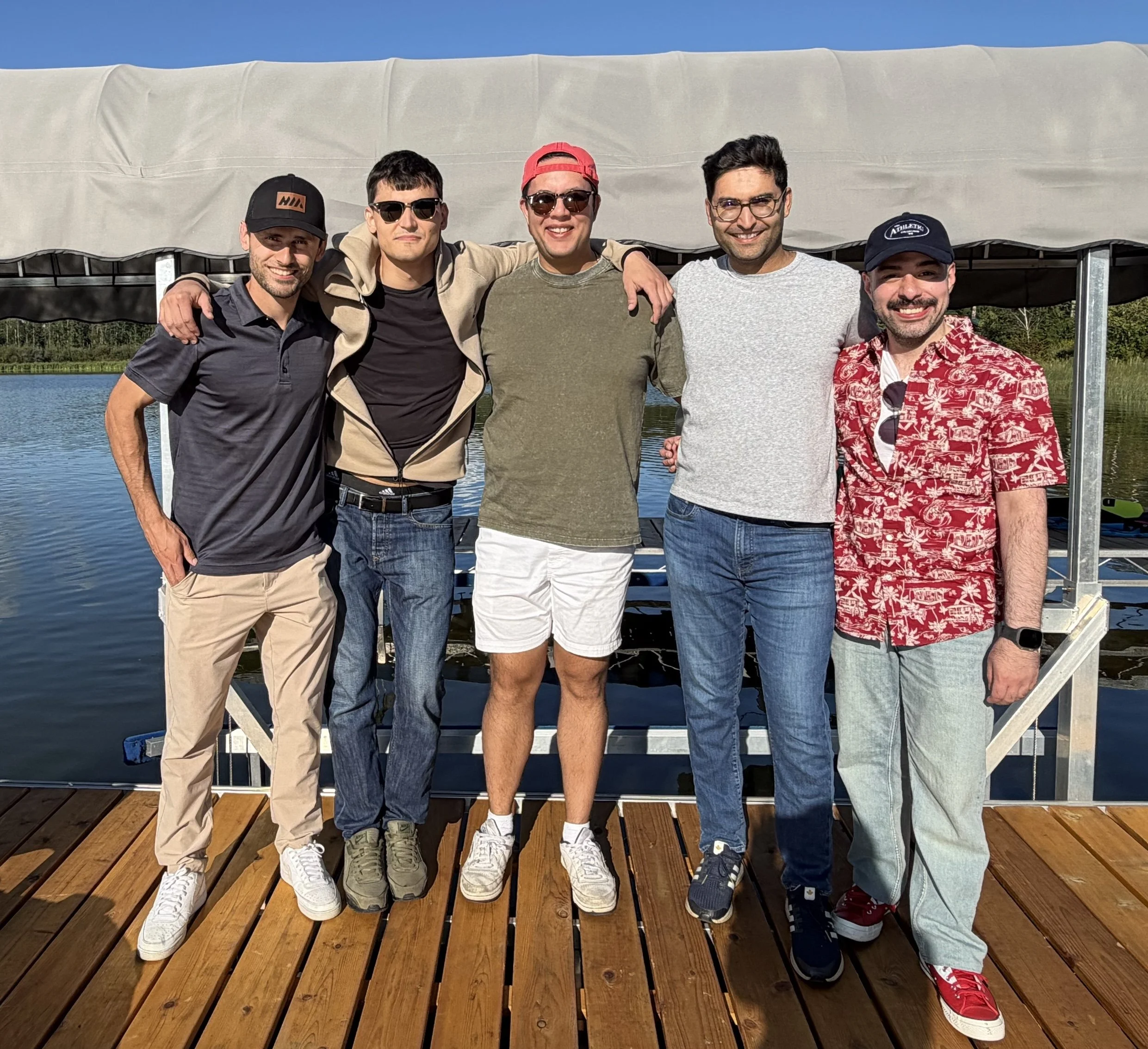 A group of five men standing arm-in-arm on a wooden dock near water, smiling, with a tent overhead and trees in the background, during a sunny day.