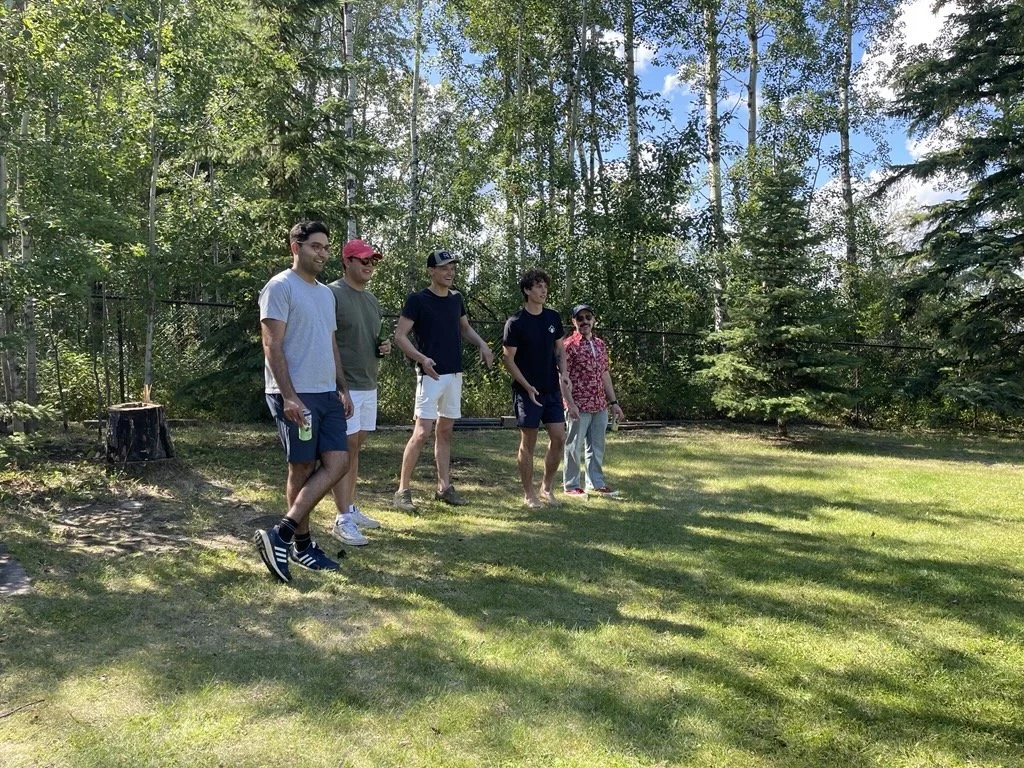 Group of five people standing on a grassy lawn surrounded by trees on a sunny day.
