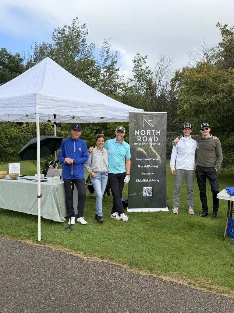 Group of five people standing outdoors on grass next to a North Road Investment Counsel sign with services listed. A white tent, with a table underneath holding bowls, is nearby. Trees and cloudy sky in the background.