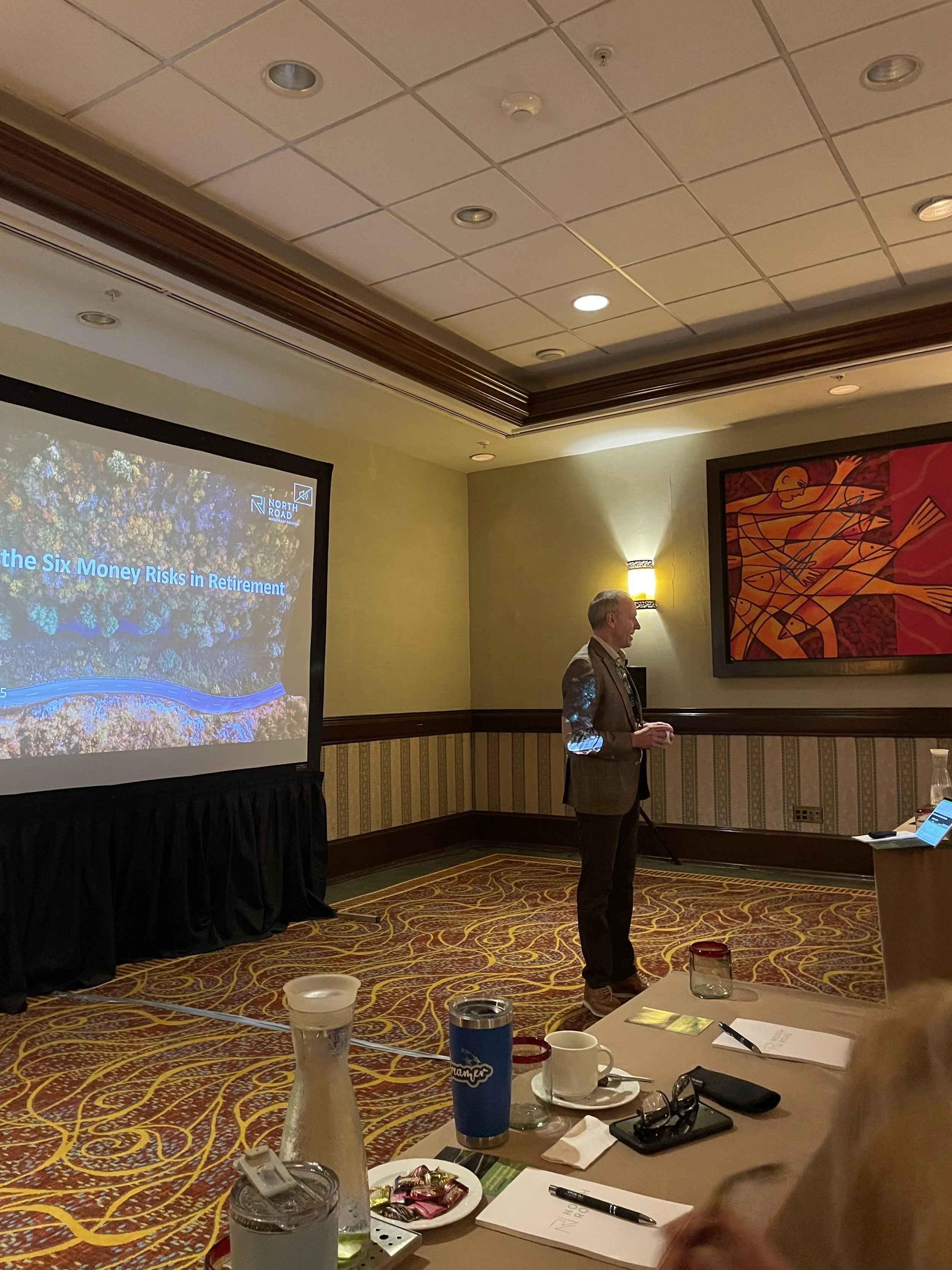 A man in a gray suit giving a presentation in a conference room. The presentation slide on the screen reads 'The Six Money Risks in Retirement.' The room has a patterned carpet, a table with various items, including coffee cups, a water pitcher, a iP